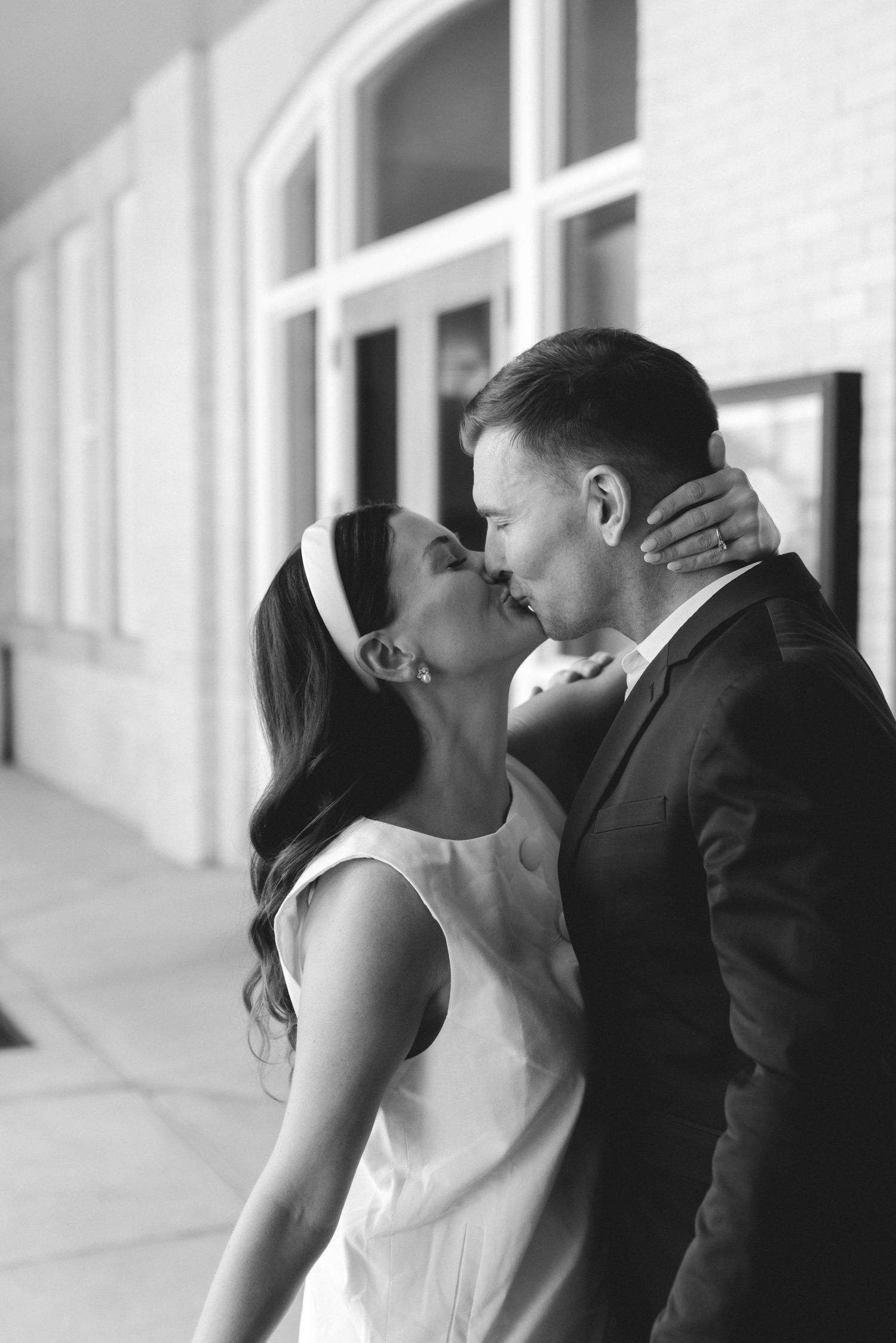 Black and white photo of a bride and groom sharing a kiss outside a building on their wedding day.