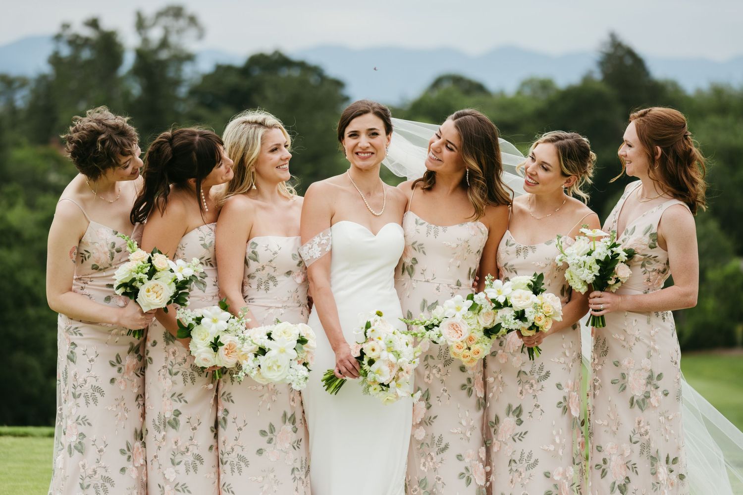 Bride in white gown poses with six bridesmaids in floral dresses holding white bouquets on a green lawn.