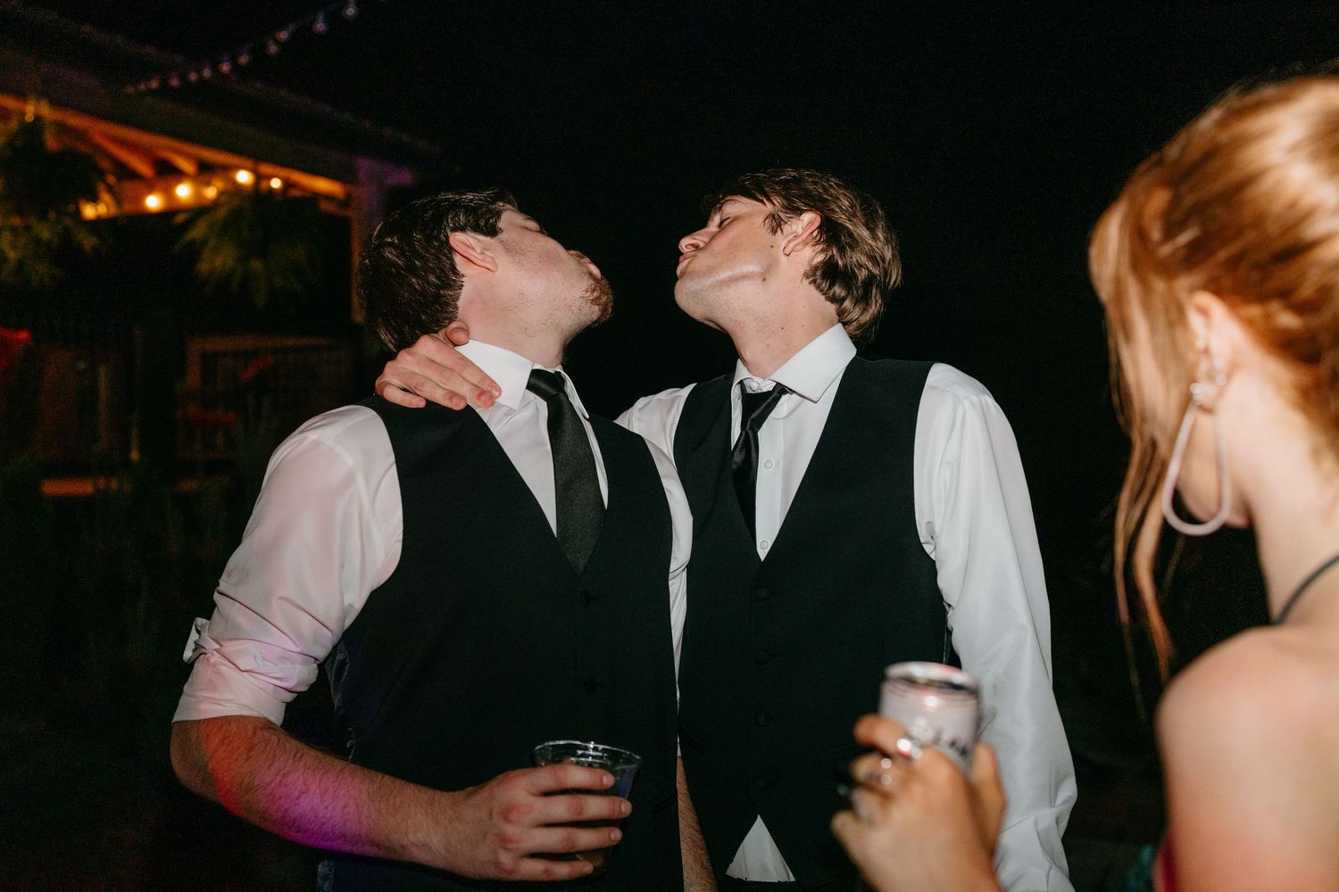 Two young men in vests and ties laugh together while holding drinks at a nighttime wedding reception.