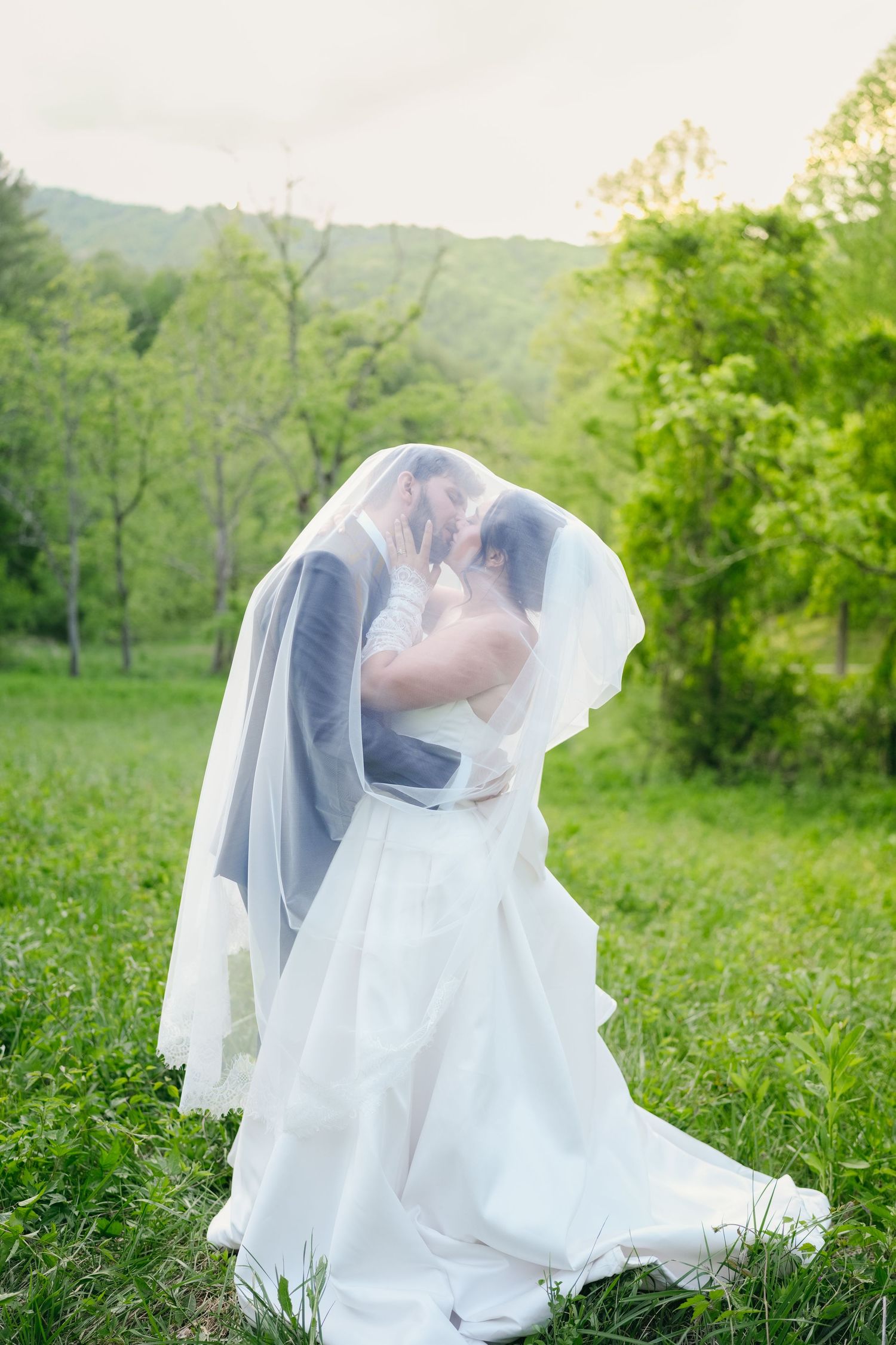 Bride and groom share an intimate kiss hidden beneath a flowing white veil in green meadow.