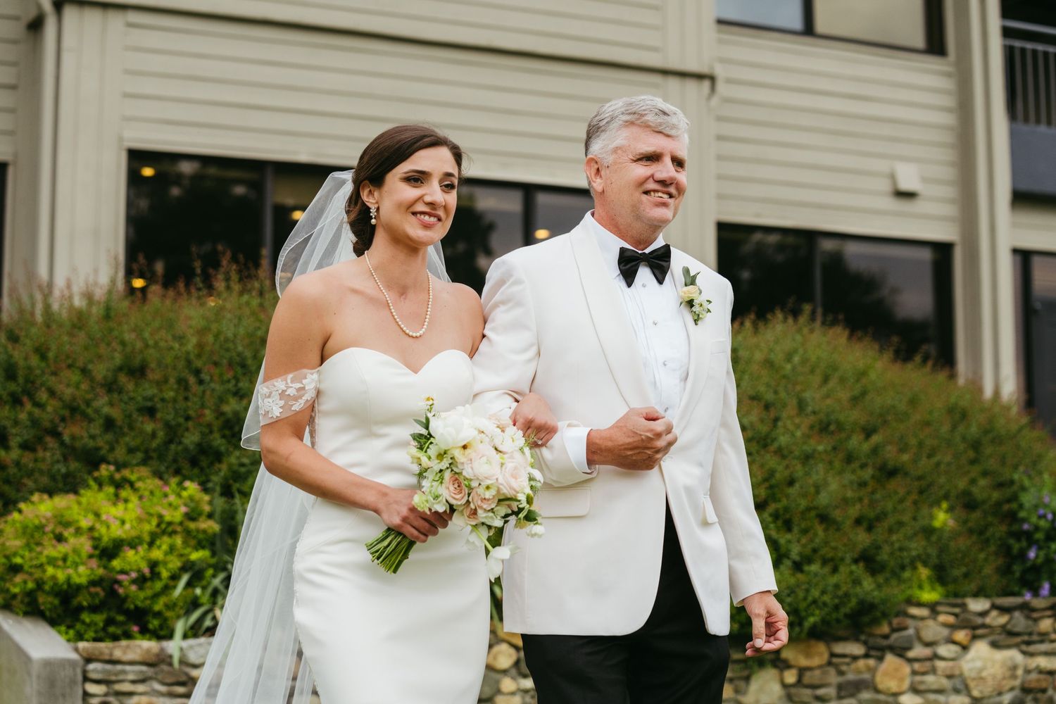 A bride in a white off-shoulder gown walks arm-in-arm with a man in a white tuxedo jacket outdoors.