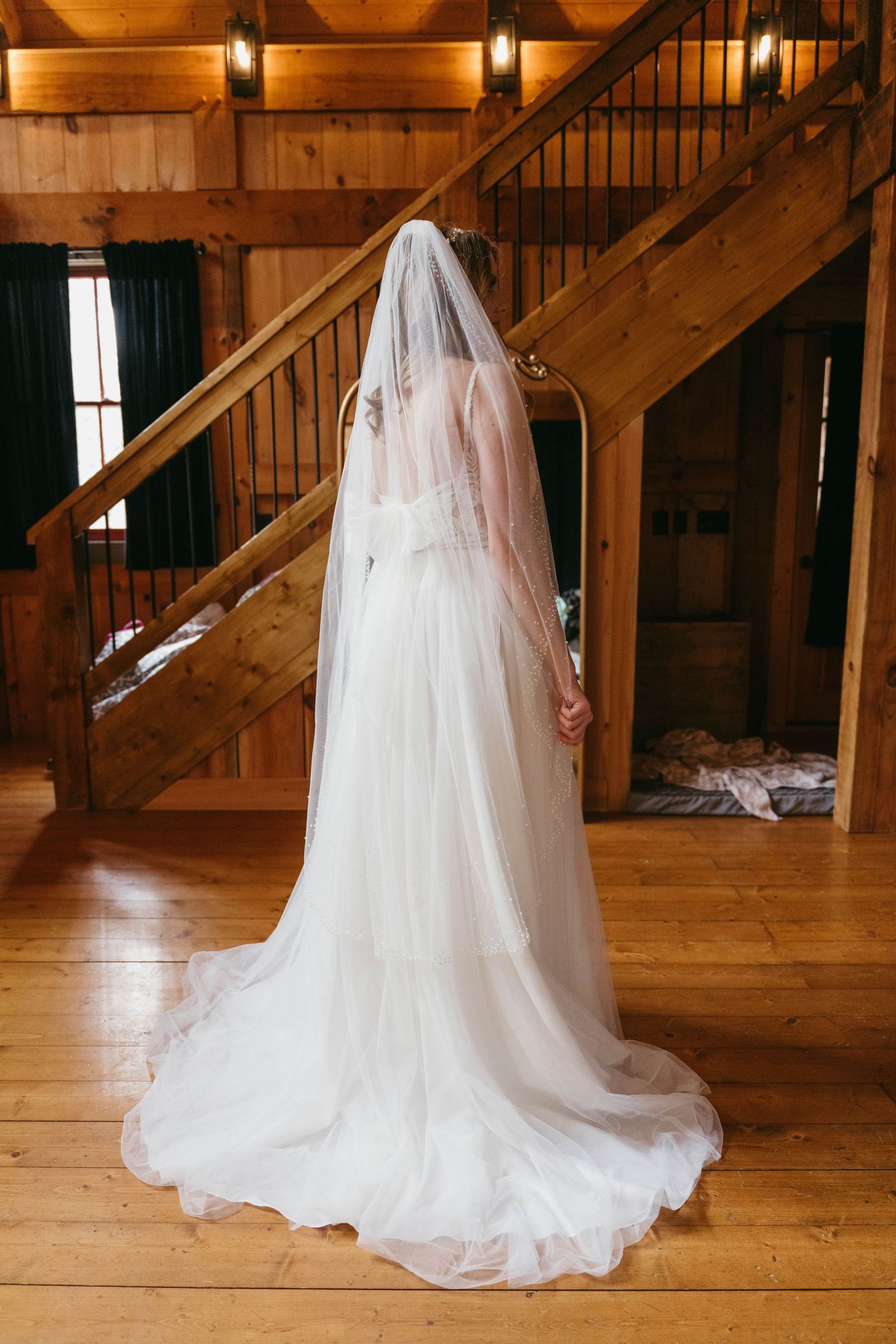 Bride in flowing white tulle wedding gown and cathedral veil standing in rustic wooden barn venue.