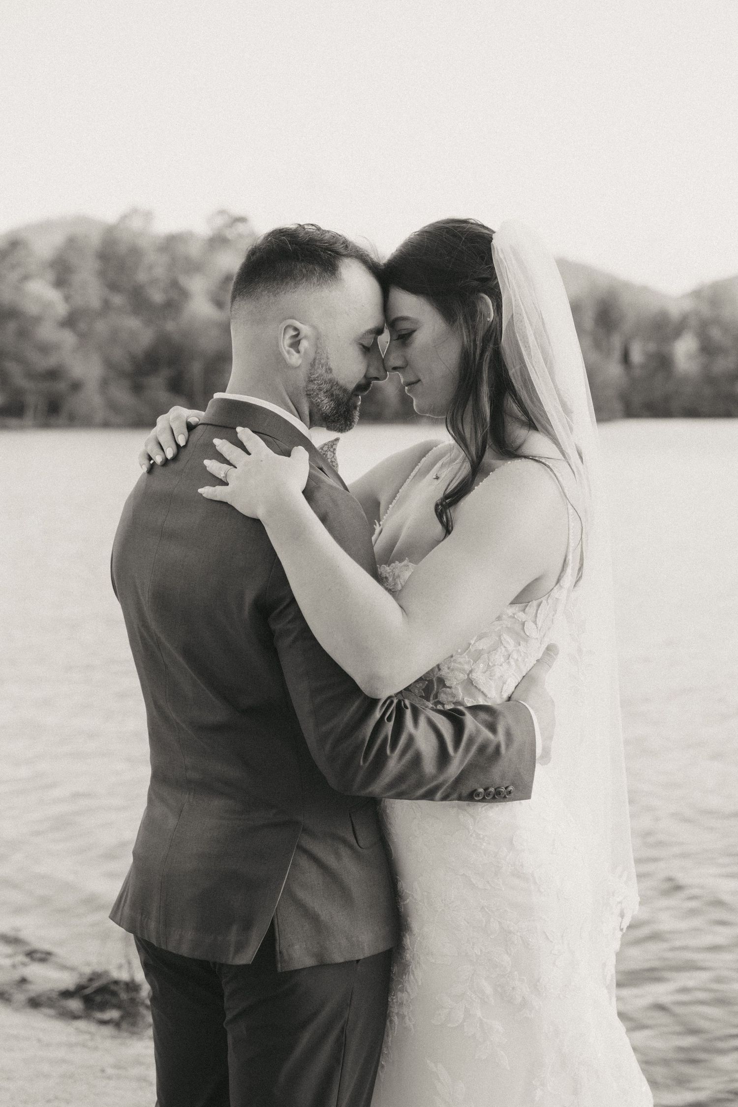 Black and white photo of bride and groom embracing tenderly by a lake.