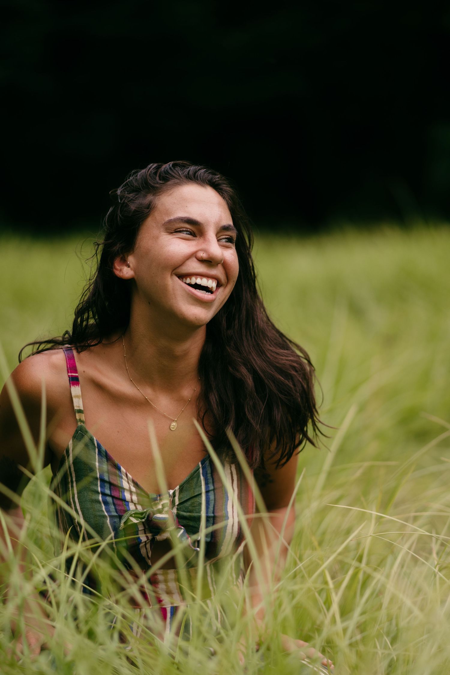 Young woman with dark hair laughing joyfully while sitting in tall green grass outdoors.