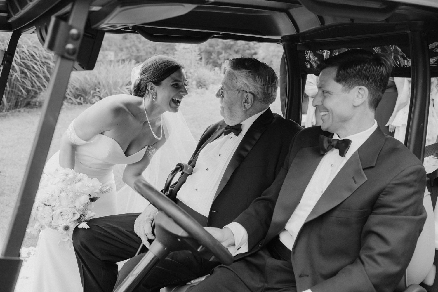 A bride and groom share a joyful moment with an older gentleman while riding in a golf cart, in black and white.