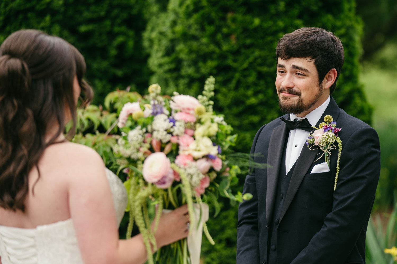 Groom in tuxedo smiling at bride holding colorful floral bouquet during outdoor ceremony.