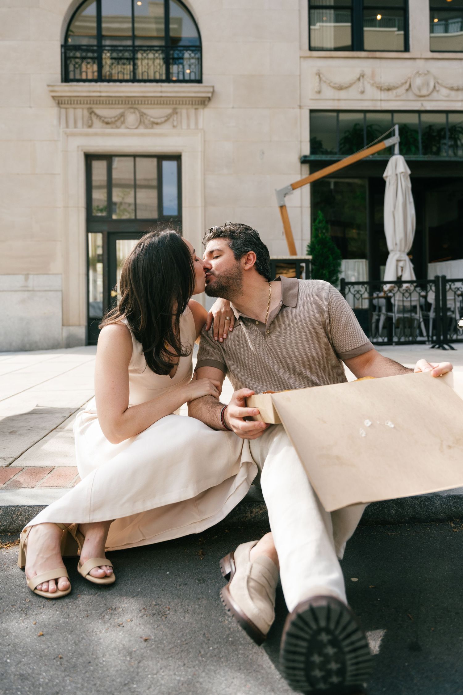 Romantic couple sharing a kiss outdoors near a classic building, sitting together with shopping bags on a sunny day.