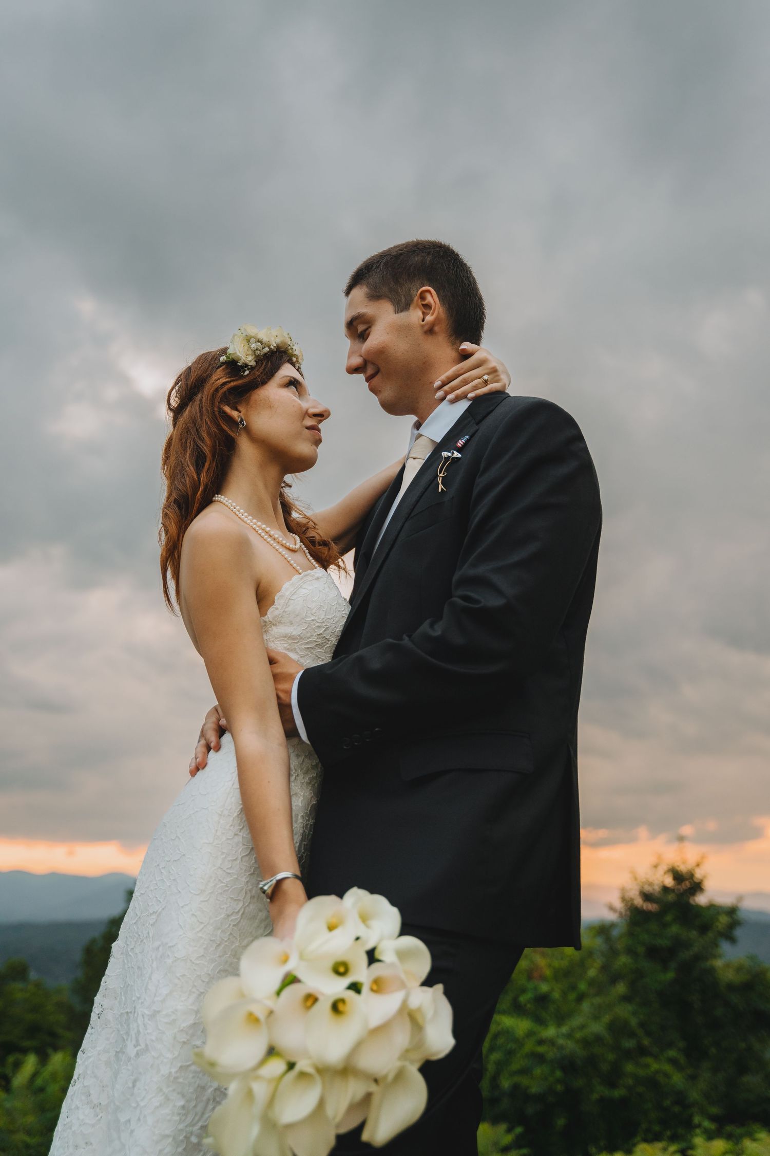 Bride and groom embrace outdoors at sunset, she holds white calla lily bouquet, wearing lace gown and floral crown.
