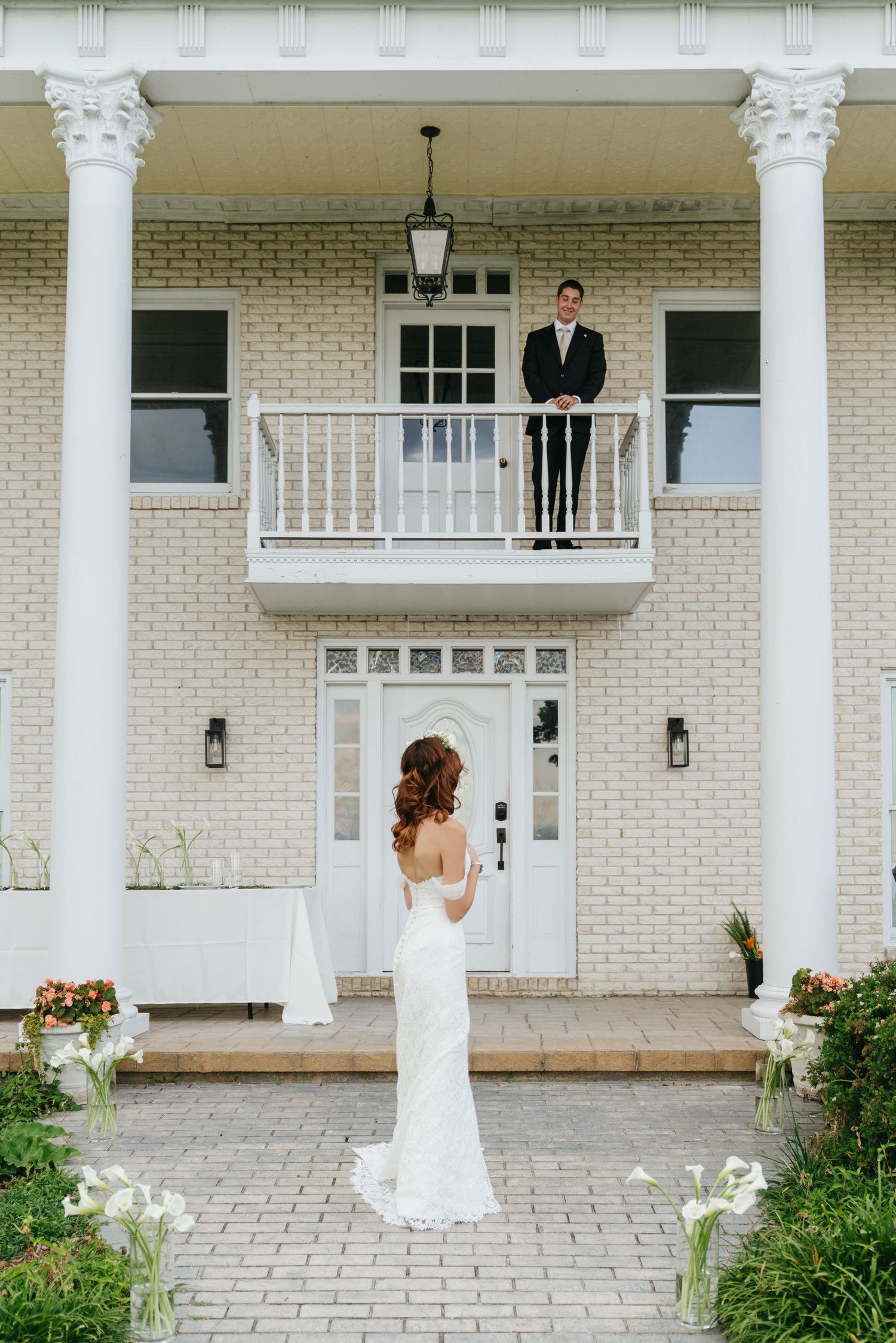 Bride in white gown stands before elegant white brick mansion as groom watches from balcony.