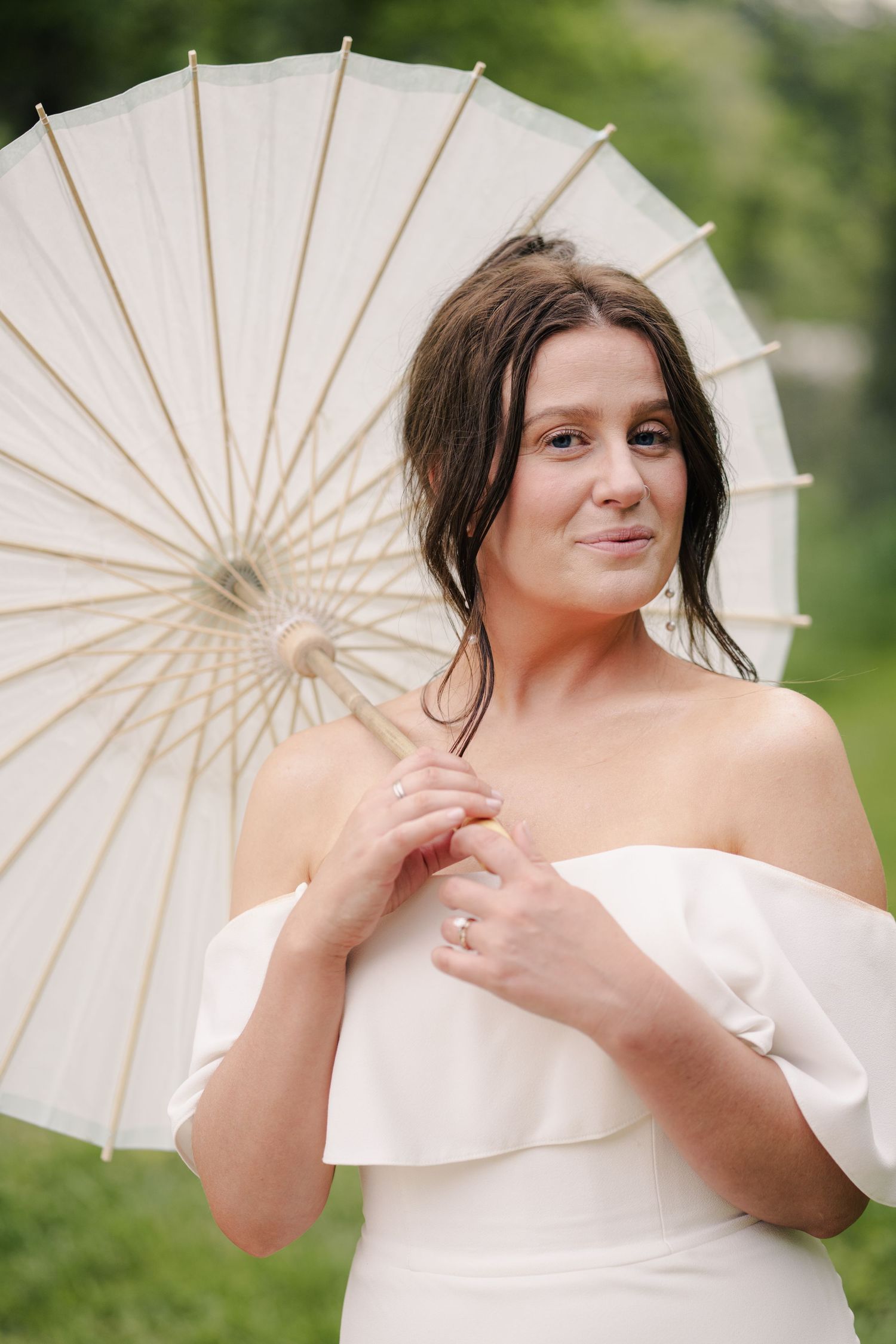 Bride in off-shoulder white gown holding white parasol umbrella outdoors.