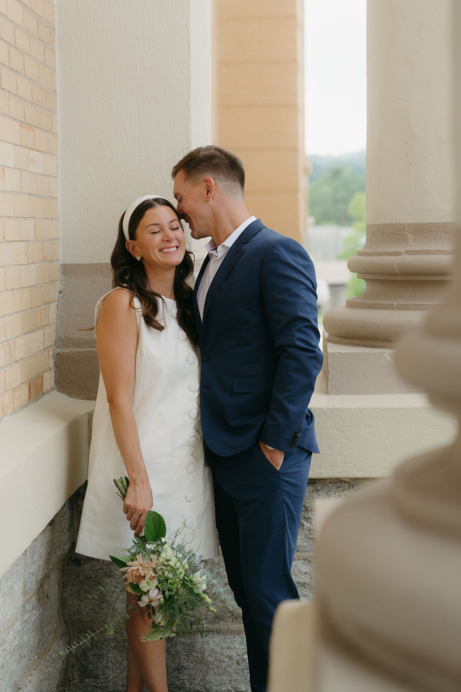 Groom in navy suit whispers to smiling bride in short white dress beside stone columns.