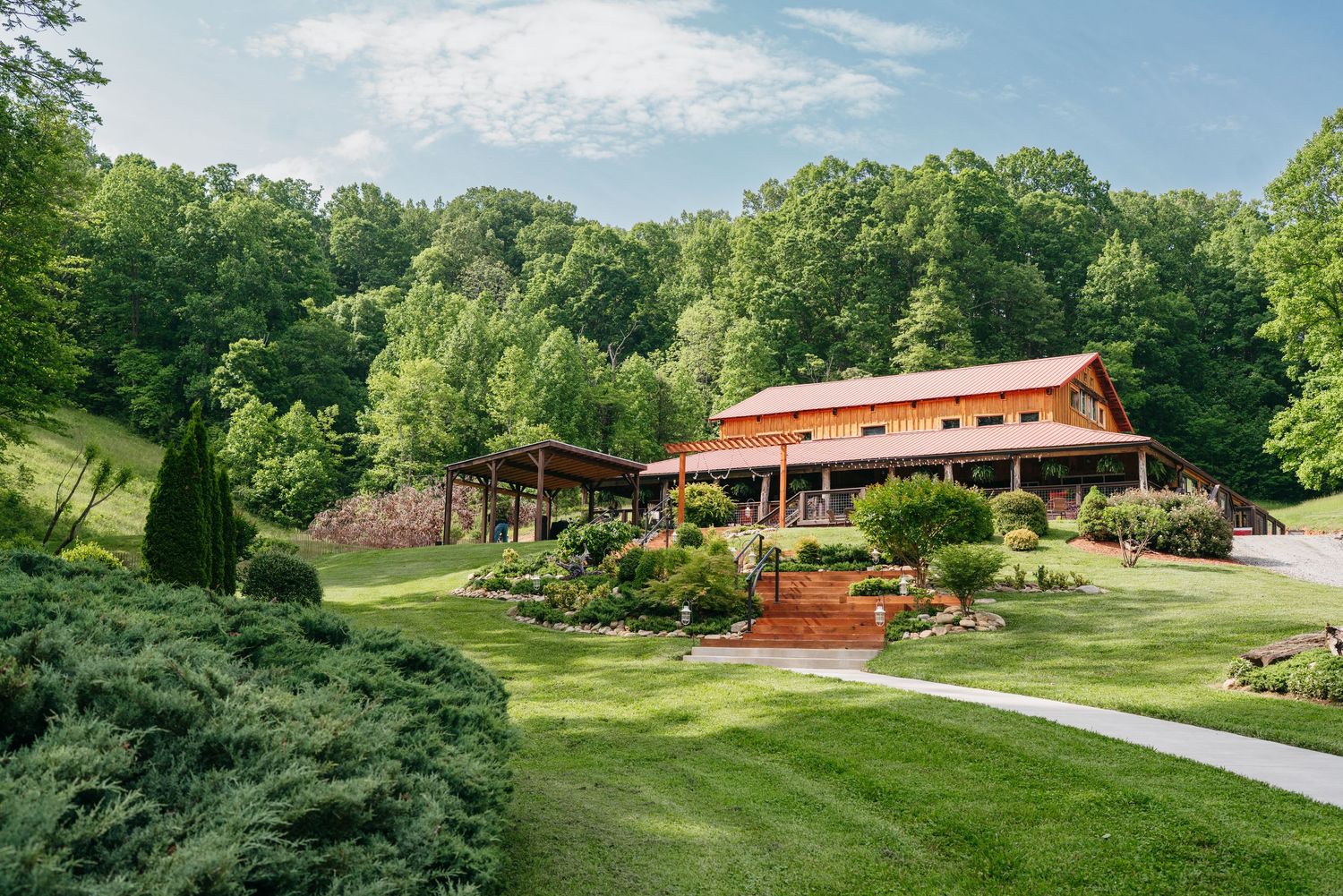 Rustic barn wedding venue surrounded by lush green trees and manicured gardens in a scenic mountain valley.