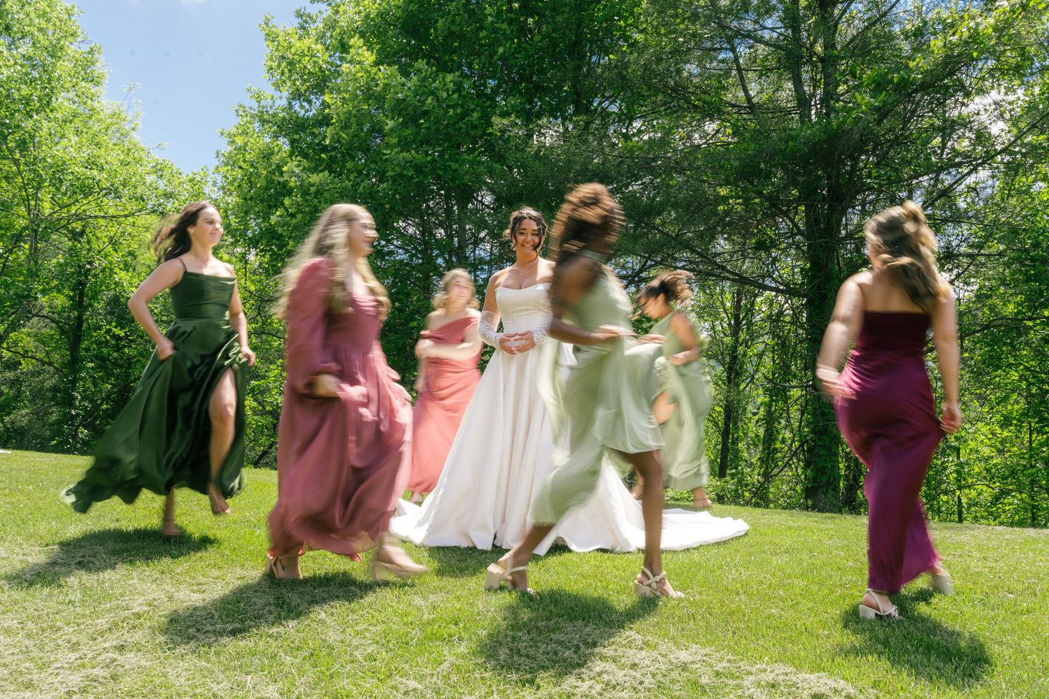 Bride celebrates with colorful bridesmaids dancing on a sunny green lawn with trees.