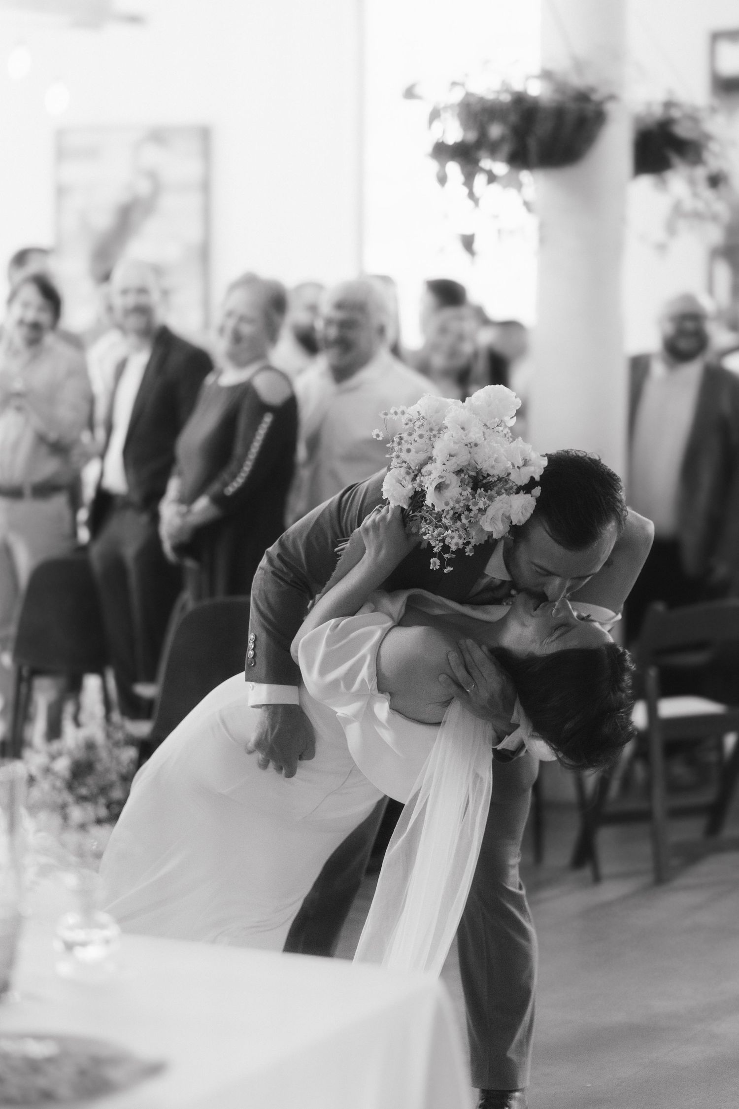 Groom dipping bride for a romantic kiss while wedding guests cheer in background.