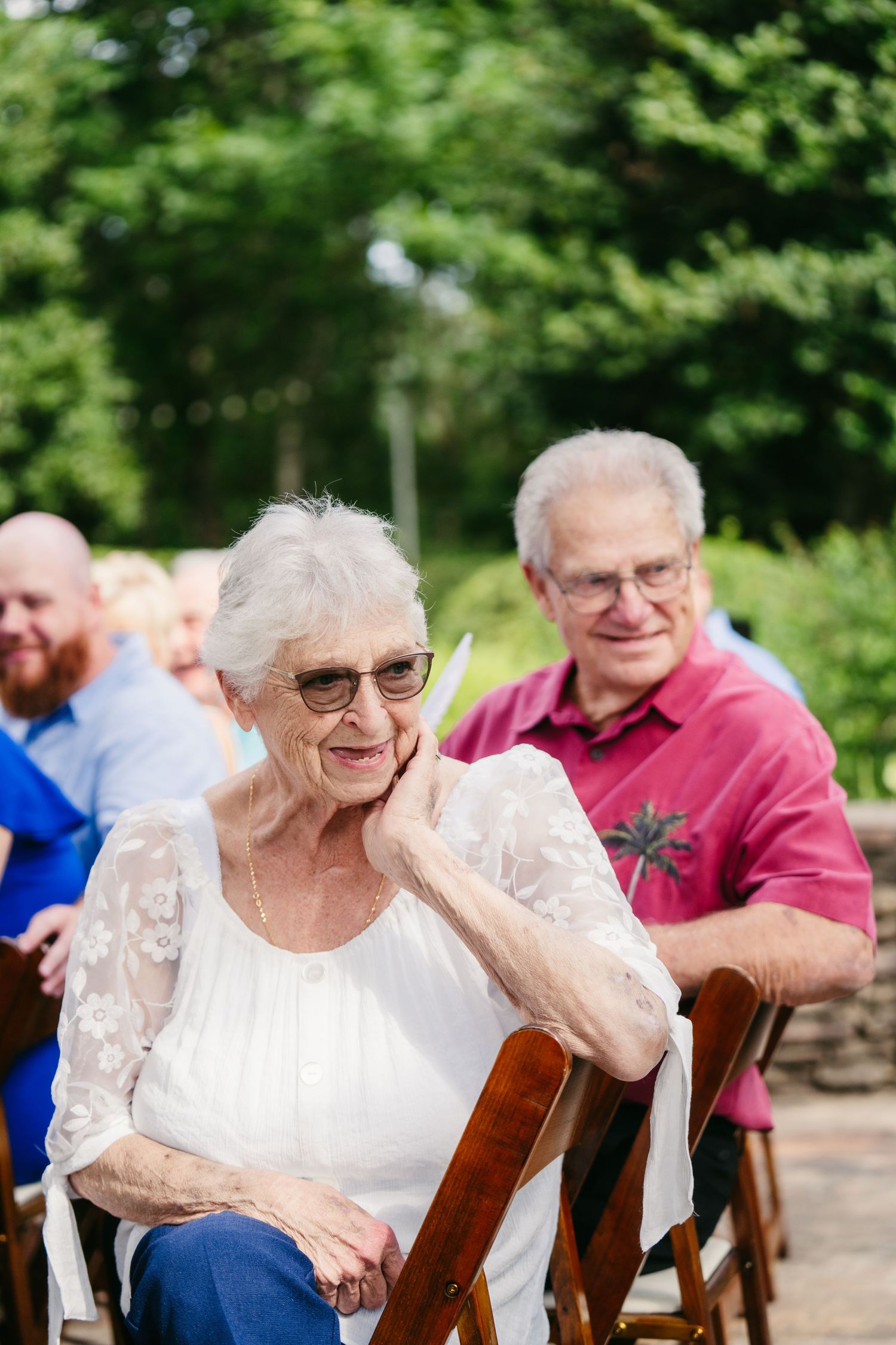 Elderly woman in white lace top smiling at outdoor wedding ceremony with guests seated behind her.
