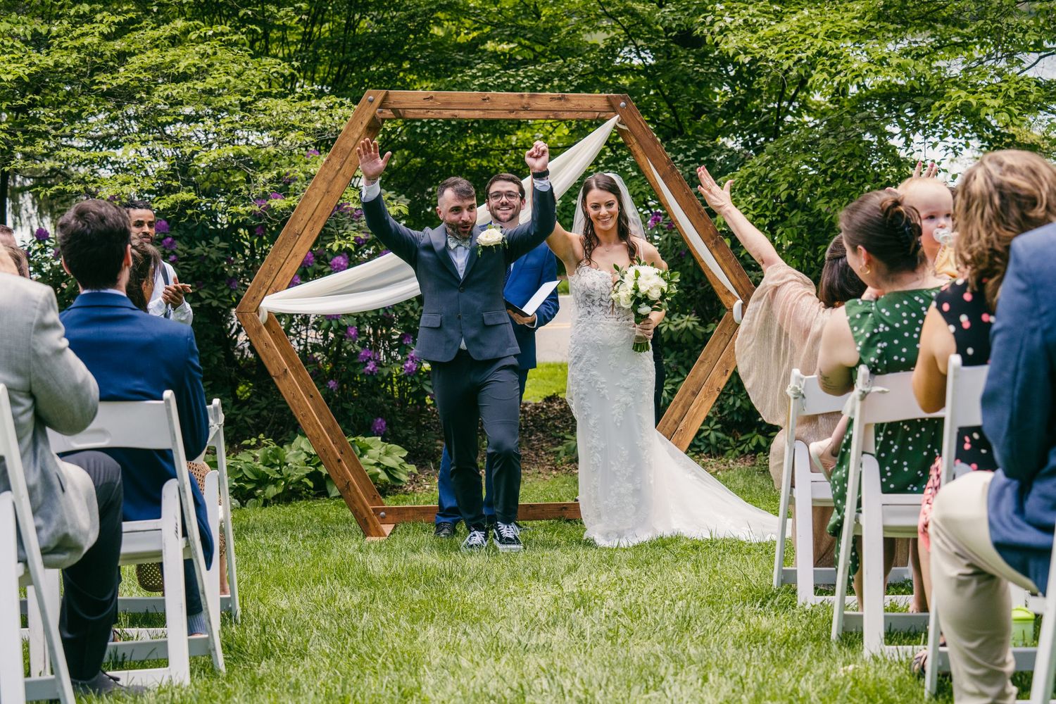 Joyful newlyweds celebrate under a hexagonal wooden arch at an outdoor wedding ceremony with seated guests.