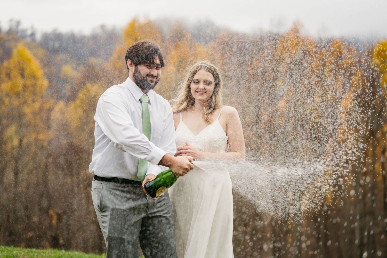 Laughing bride and groom spraying champagne bottle outdoors with vibrant autumn foliage background.
