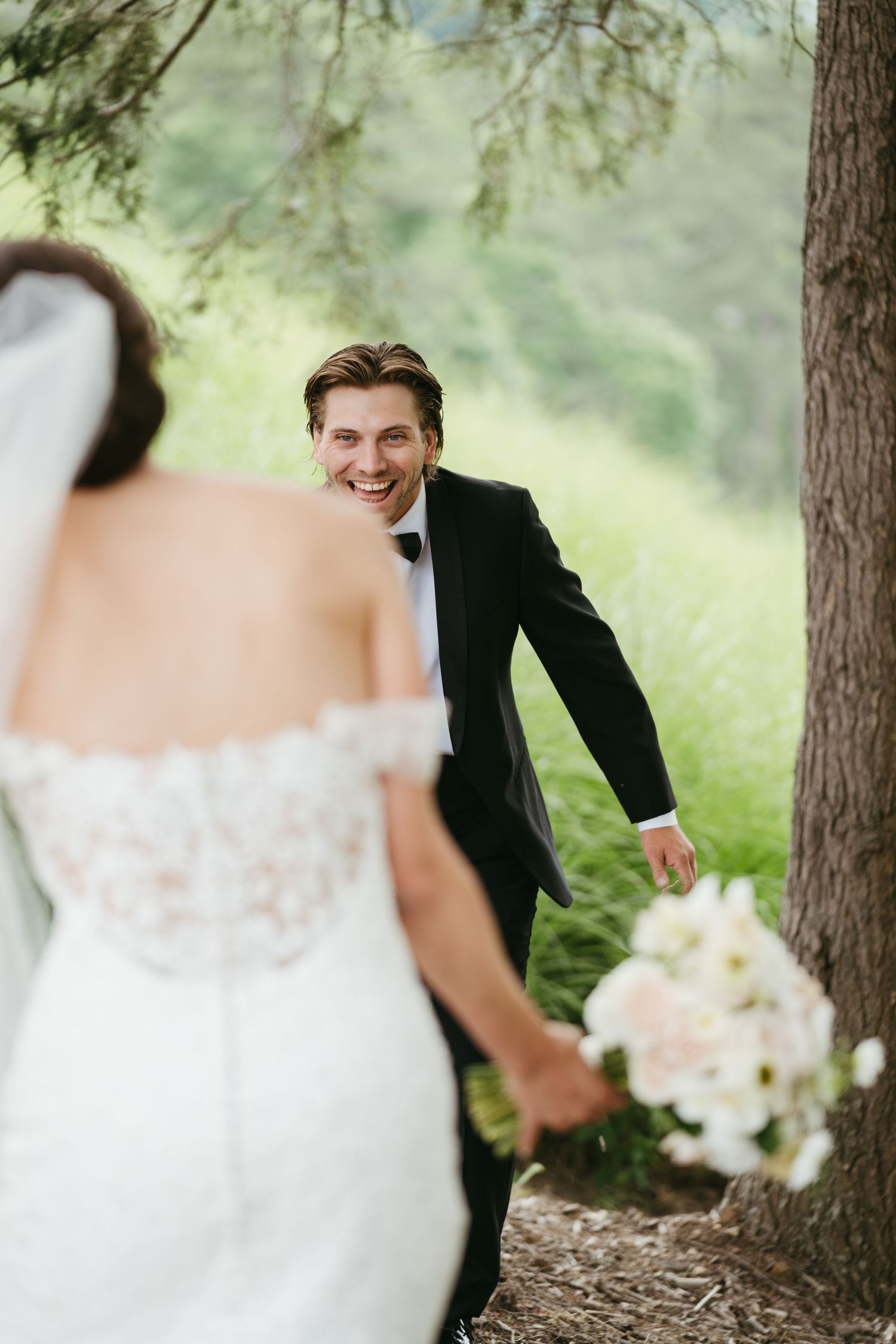 Smiling groom in black tuxedo reaches for his bride among lush green trees during wedding portraits.