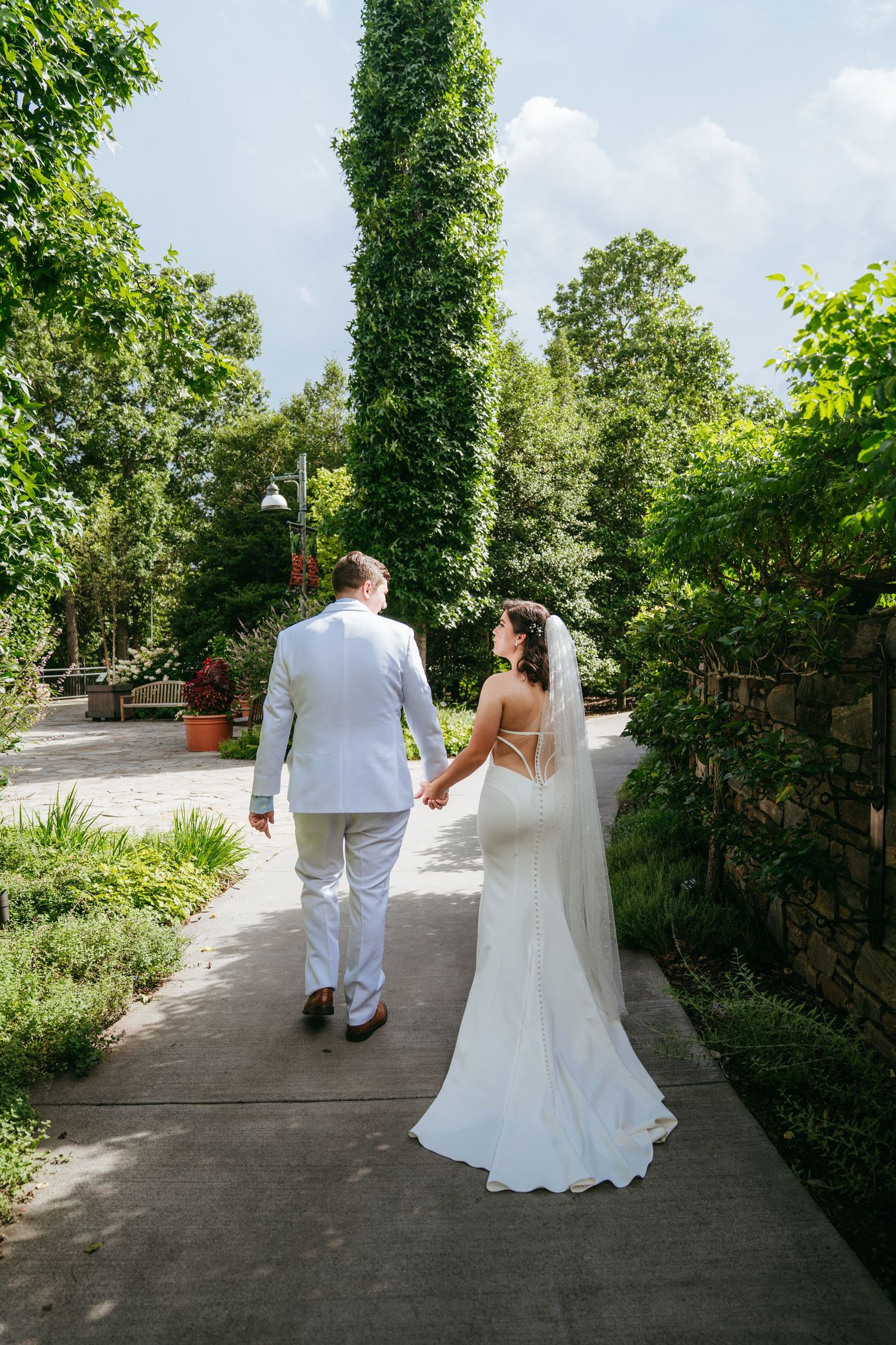 Bride and groom hold hands walking down garden path, bride wearing veil and white gown.