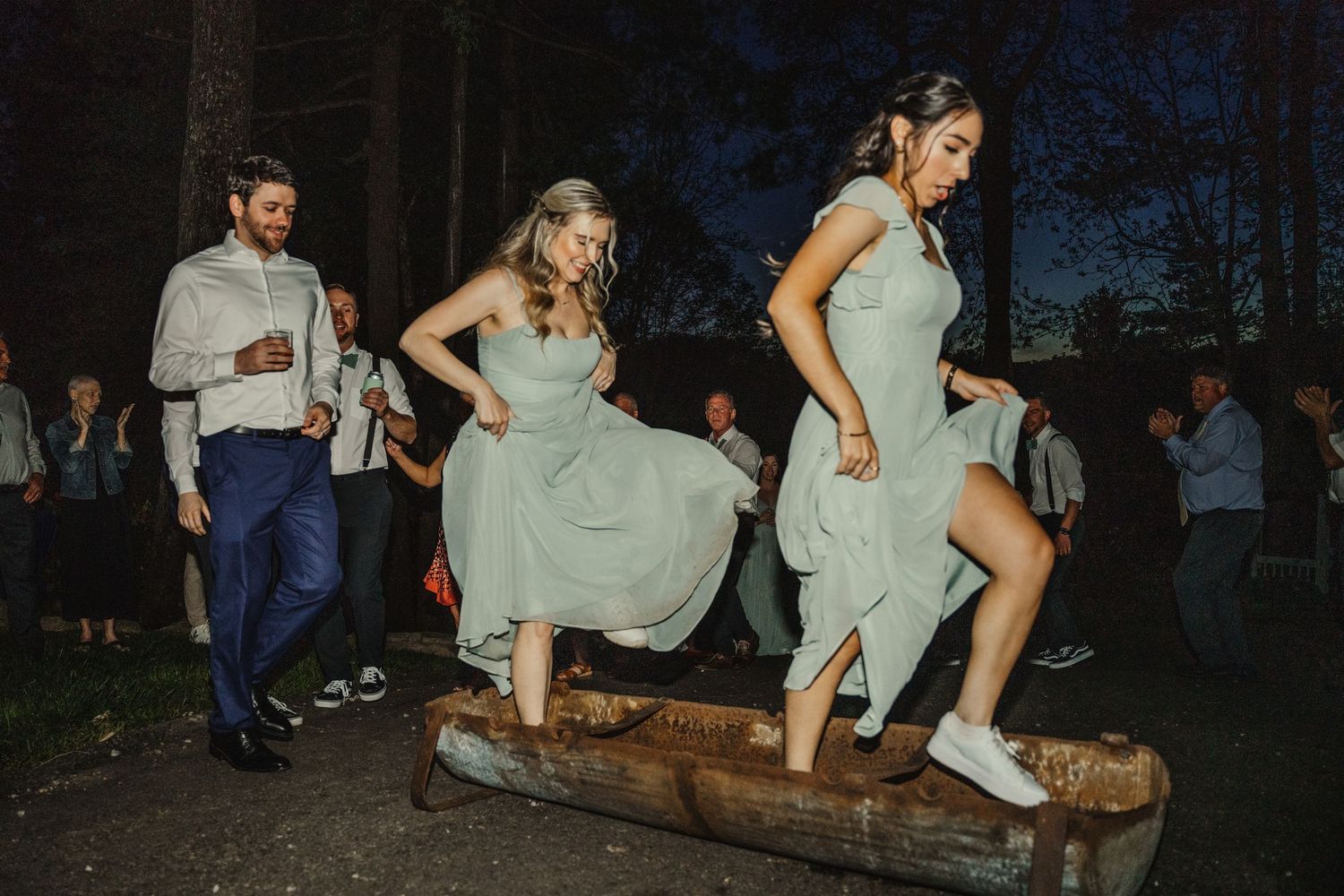 Bridesmaids in sage green dresses dance and jump over a log at an outdoor evening wedding reception.