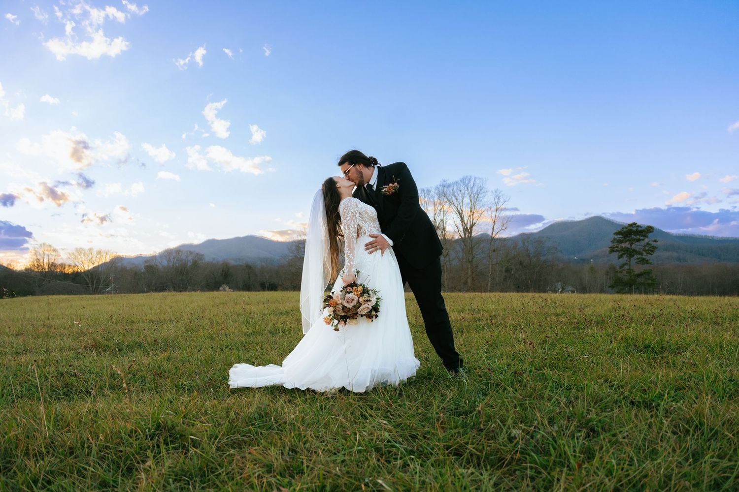 Bride and groom sharing romantic kiss in open mountain field at sunset during wedding.
