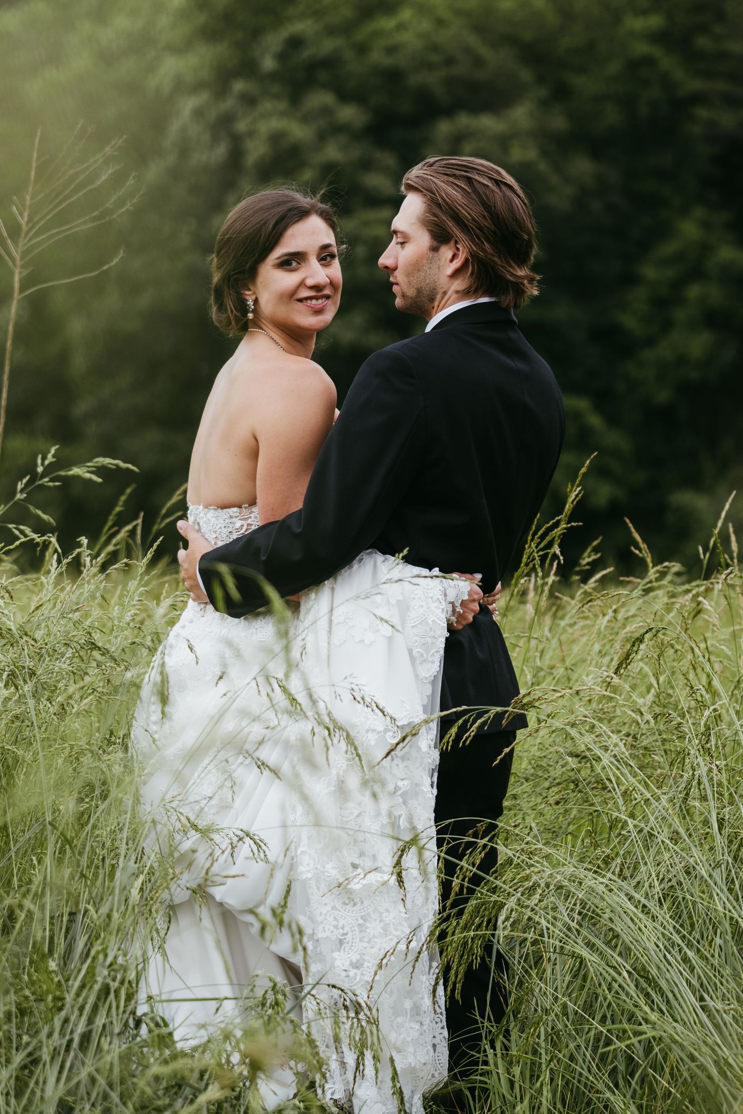 Bride in white lace gown embraces groom in black tuxedo among tall green grass in a romantic outdoor wedding portrait.