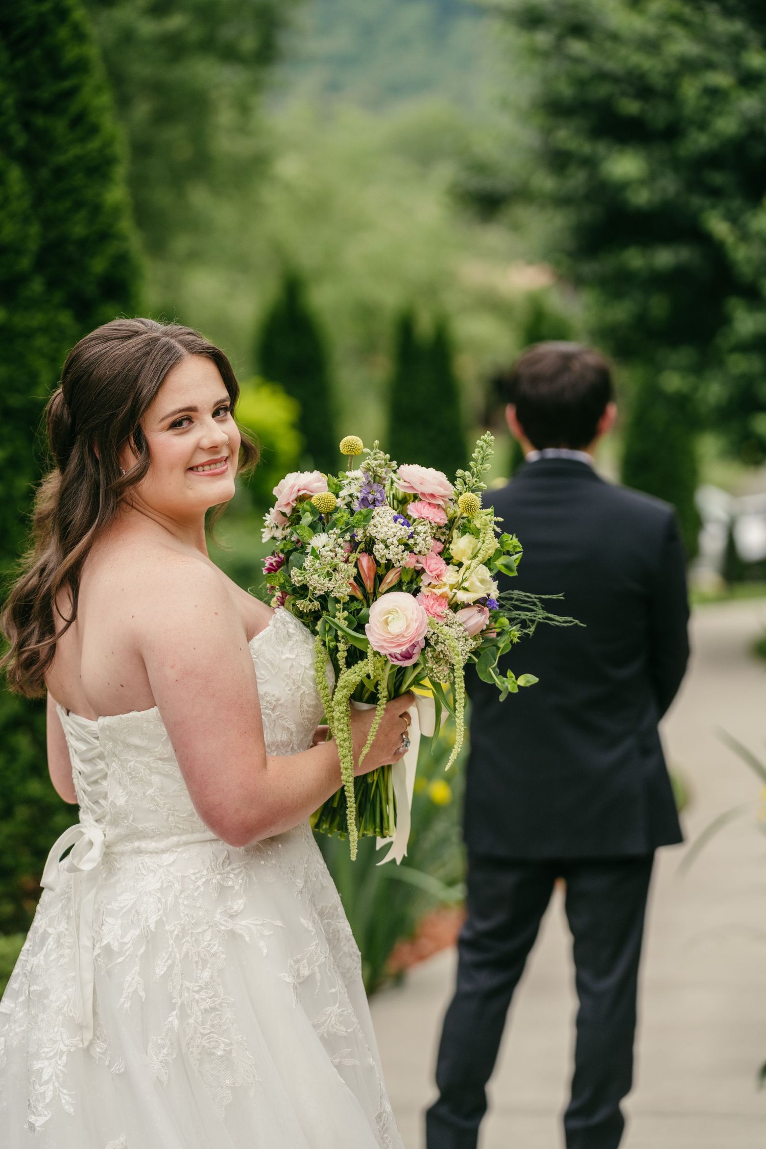 Bride in strapless lace gown holds pink bouquet while groom stands in background.