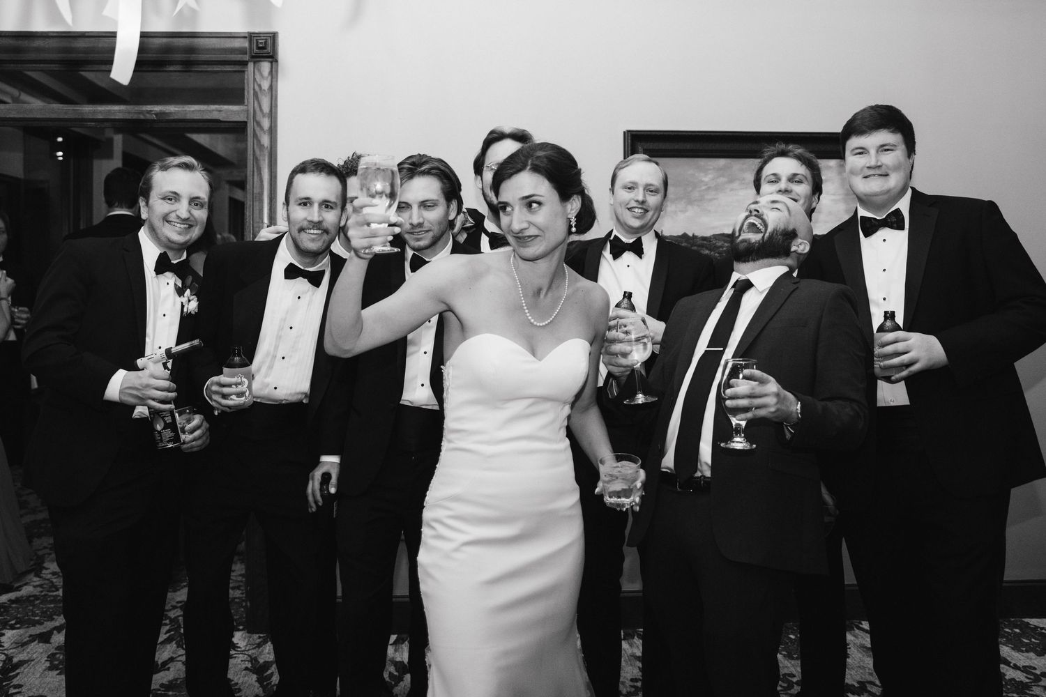 Bride in white gown poses with groomsmen in tuxedos holding champagne glasses at wedding reception.