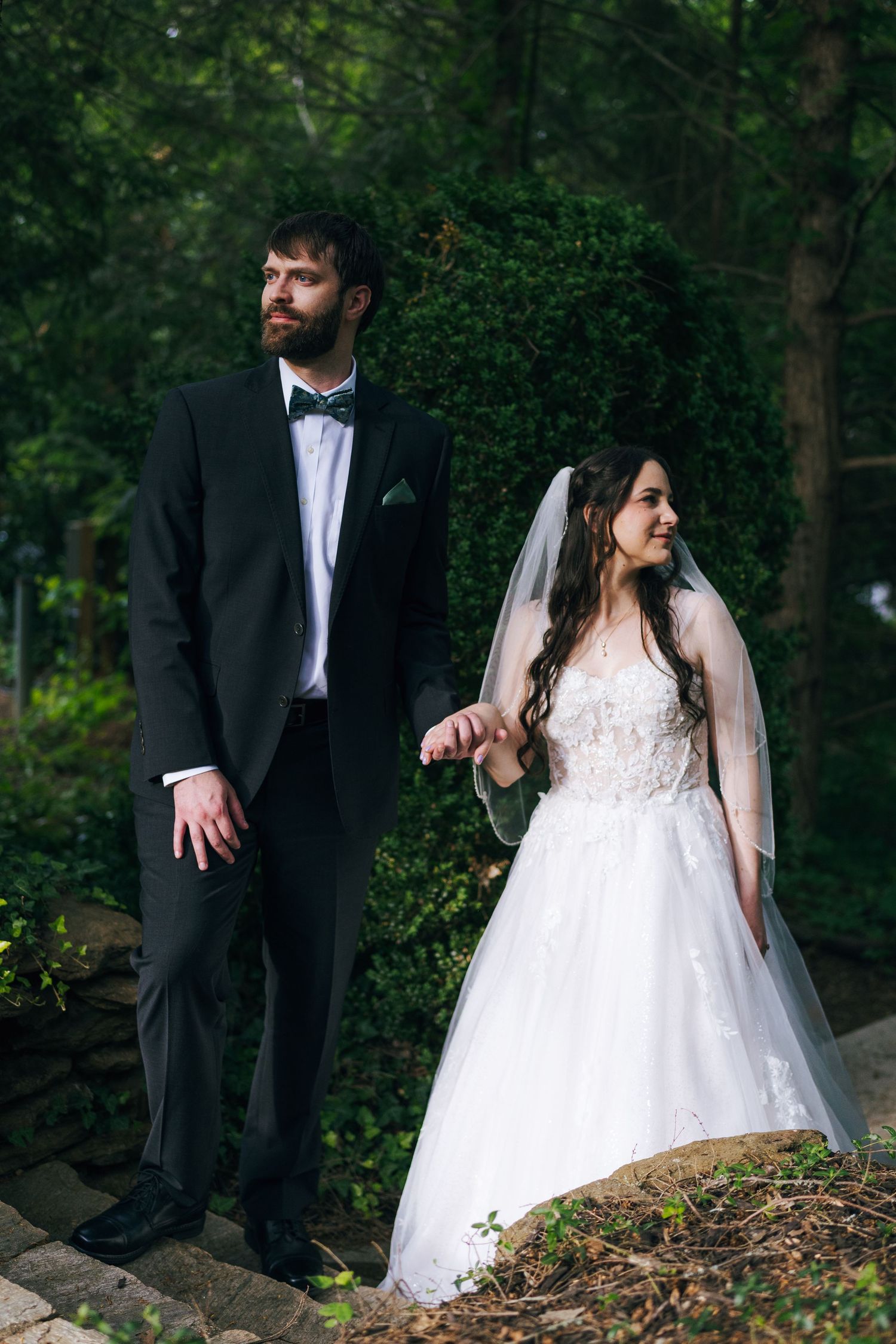 Elegant bride and groom stand together on stone steps in a lush green forest setting.