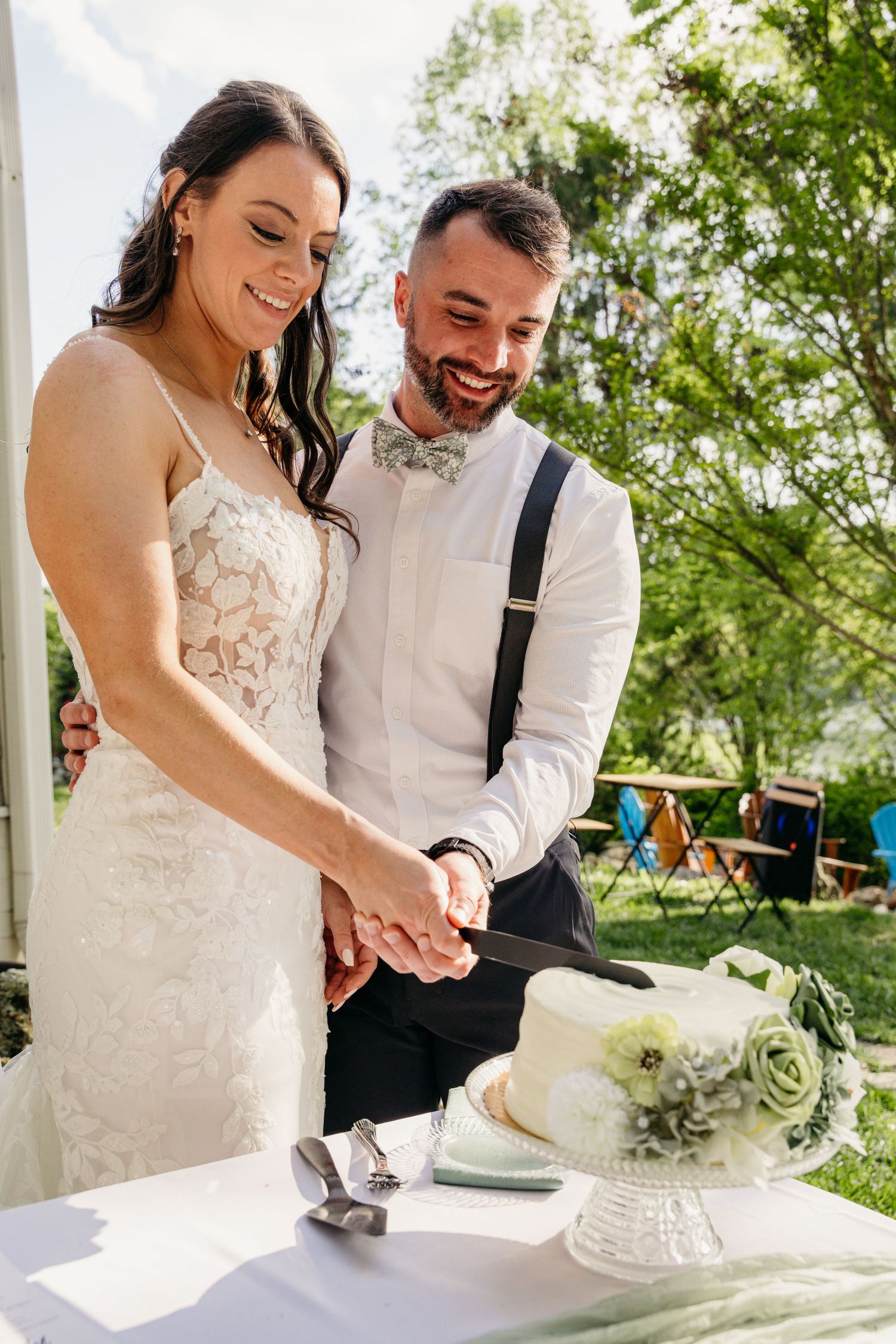 Smiling bride and groom cutting their floral-decorated white wedding cake outdoors.