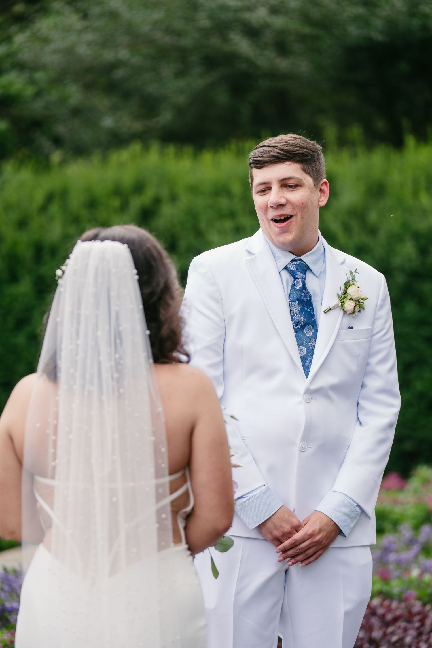Groom in white suit reacting with joy during first look with bride in garden wedding.