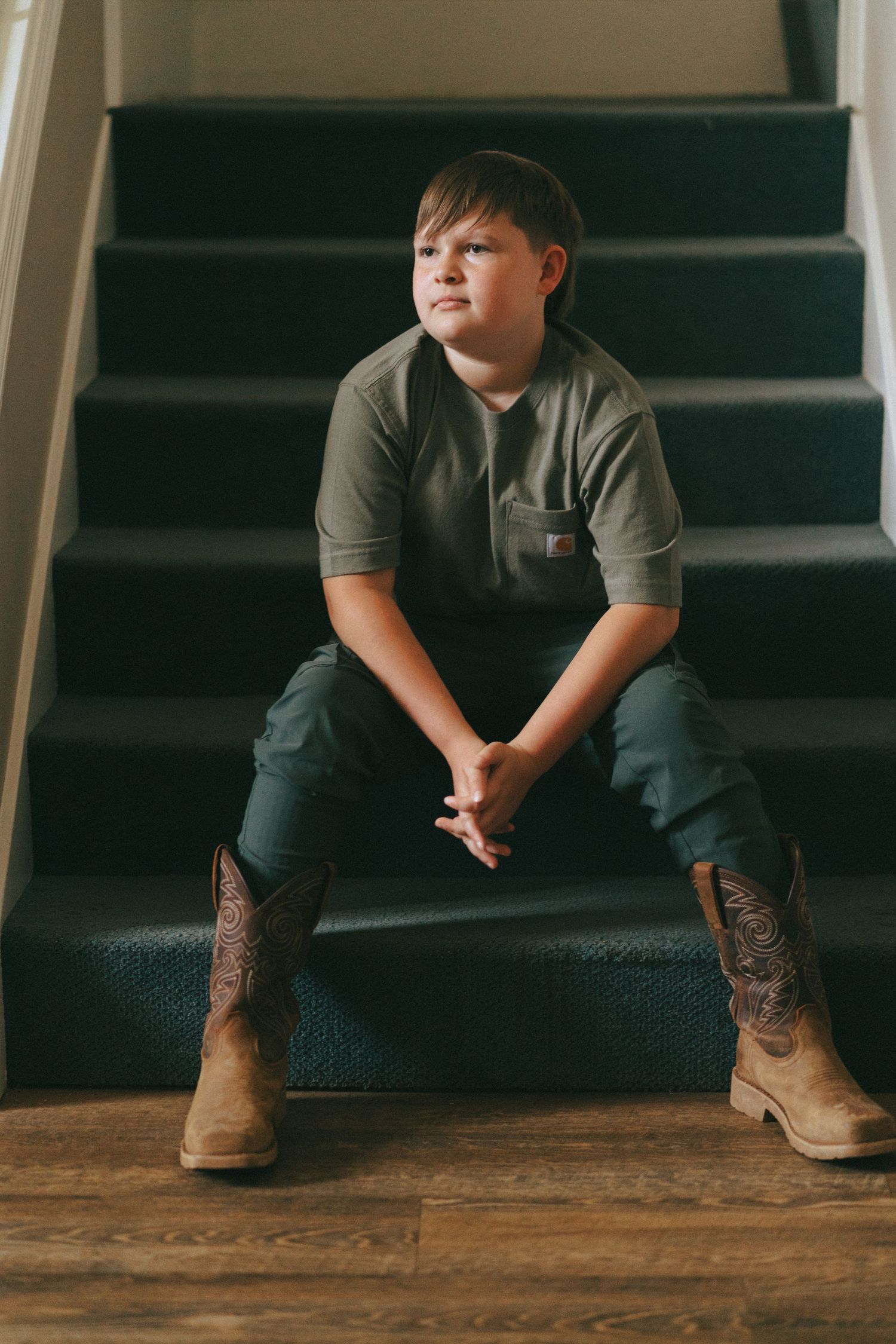 Young boy in olive shirt and cowboy boots sitting on carpeted staircase with hands clasped.