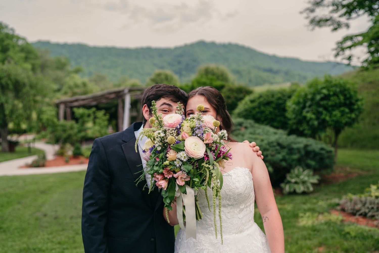 Bride and groom pose together wearing face masks while holding a lush floral bouquet with mountain views behind.