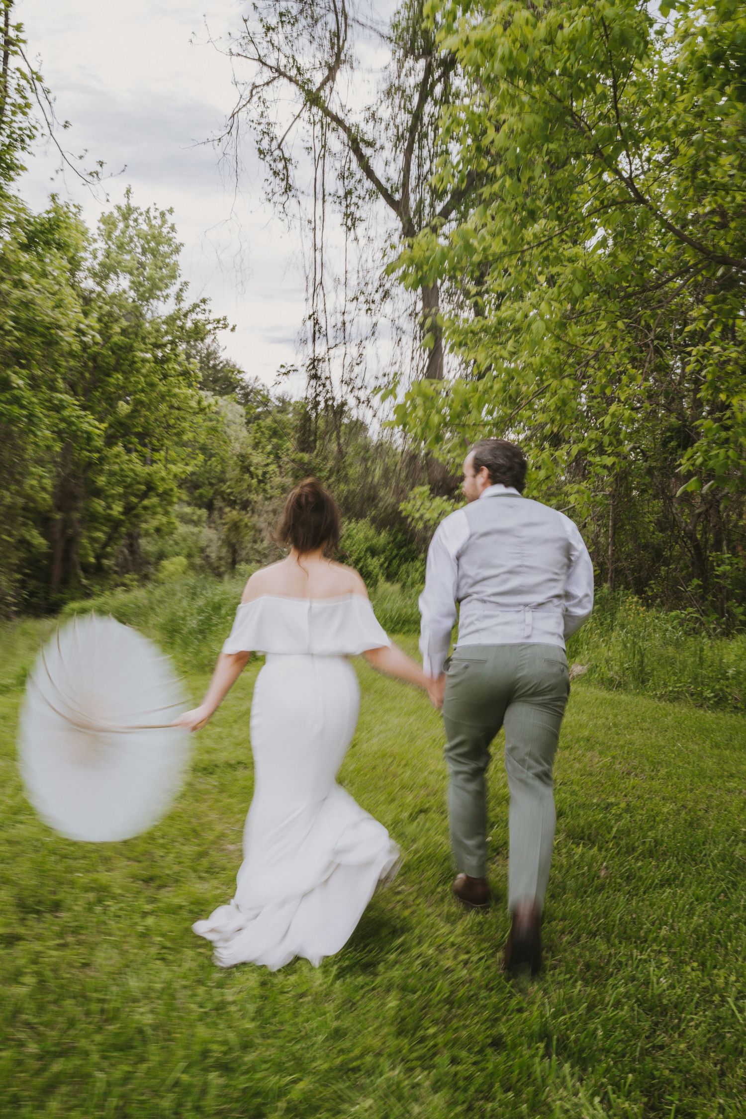 Bride and groom holding hands running through lush green outdoor landscape.