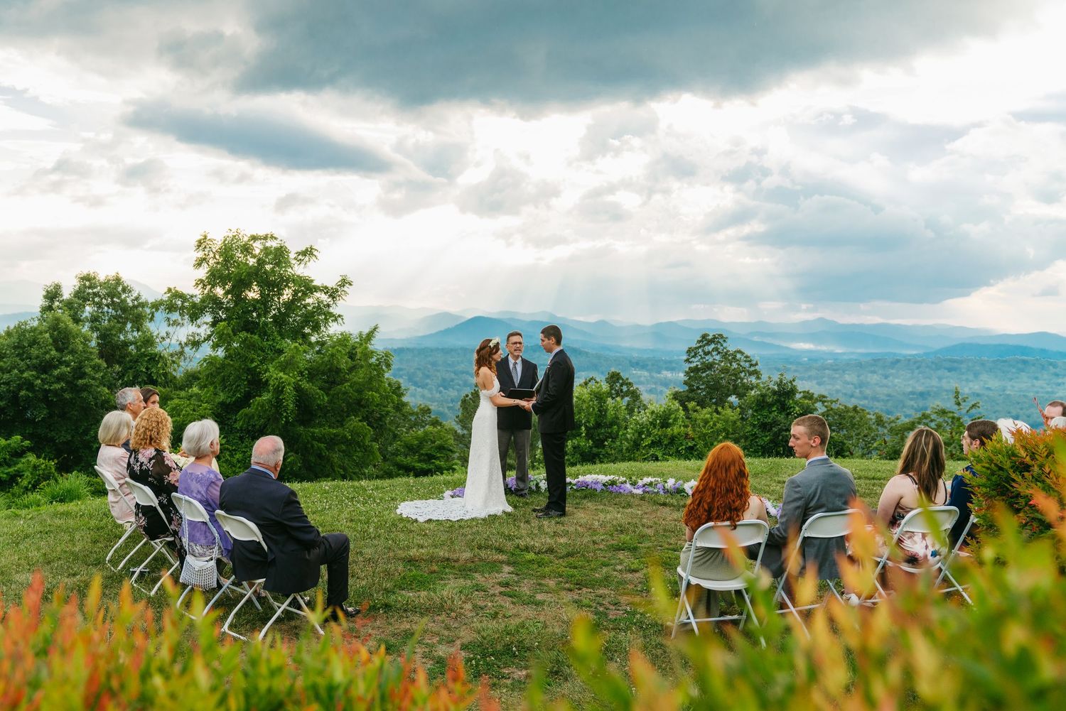 Intimate outdoor mountain wedding ceremony with small guest gathering under dramatic cloudy sky.