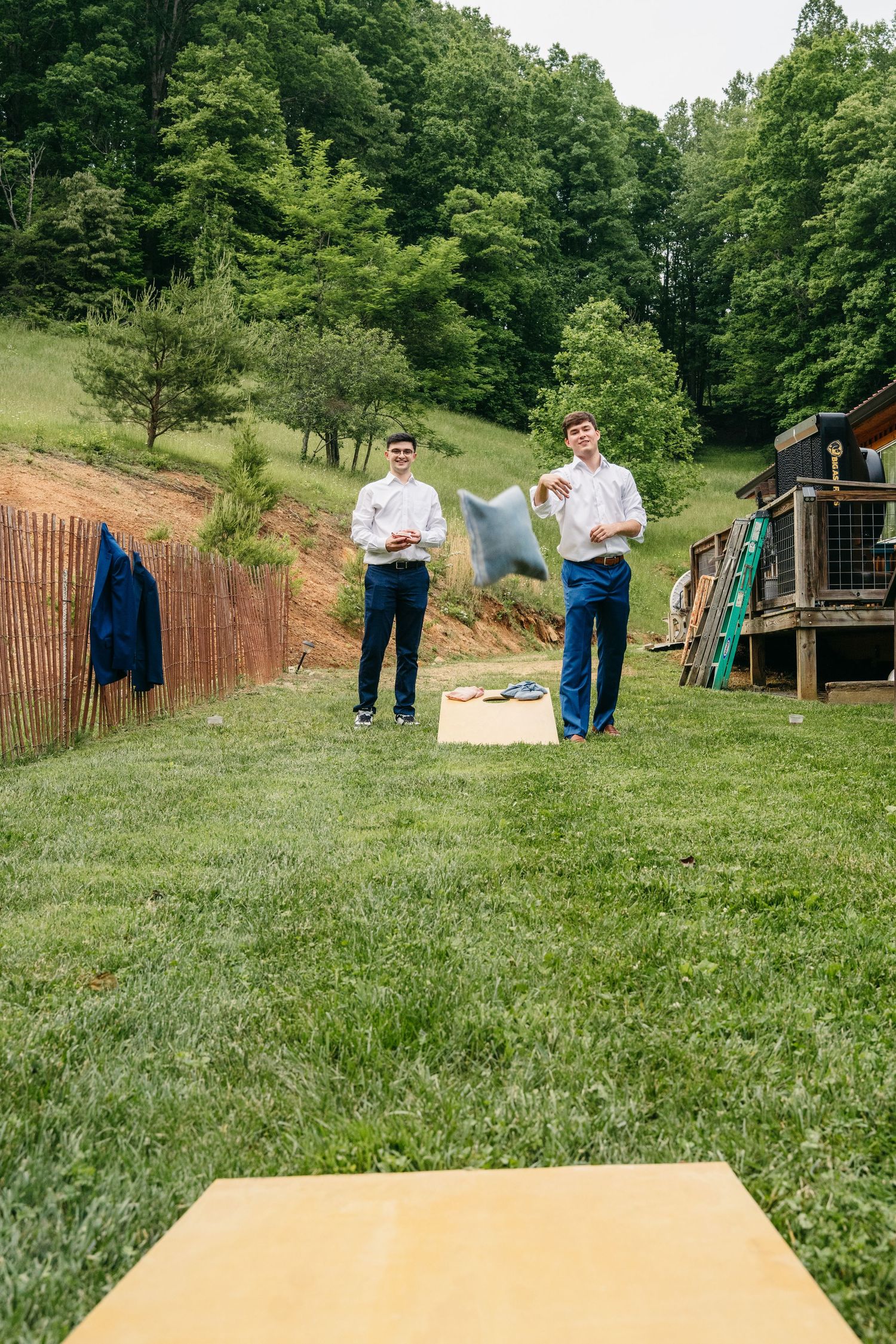 Two groomsmen in white shirts play cornhole on a green lawn with wooded hills in the background.