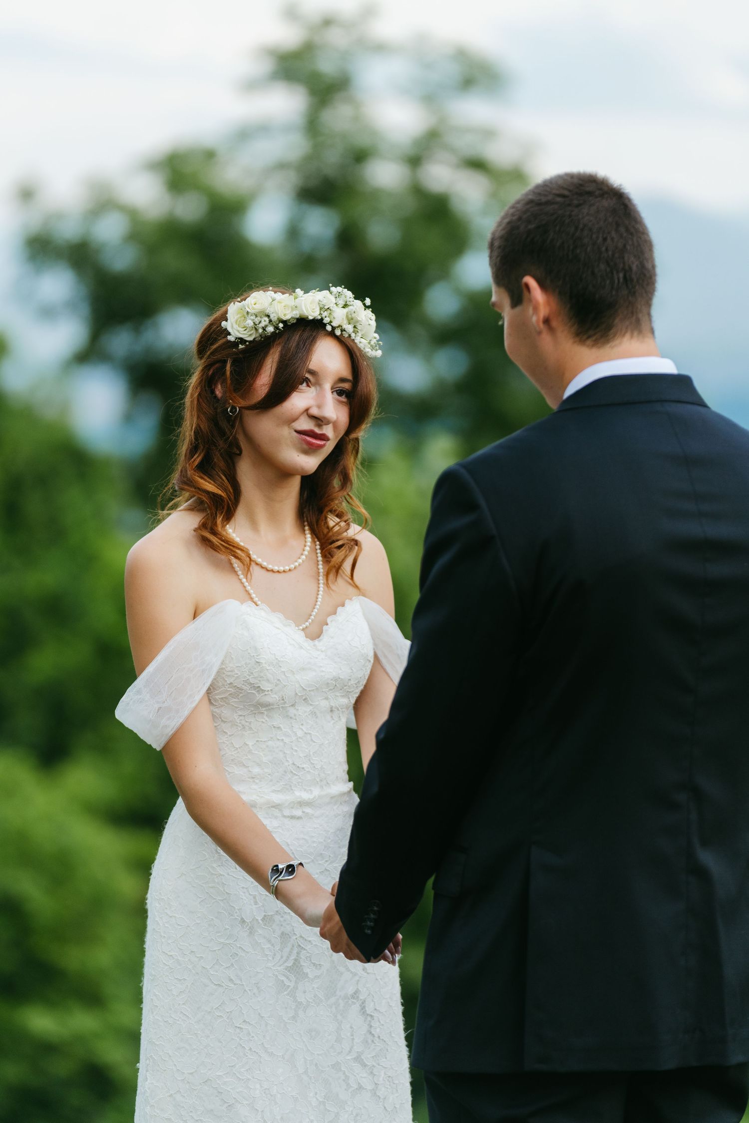 Bride wearing floral crown in lace dress holds grooms hands during outdoor mountain ceremony.