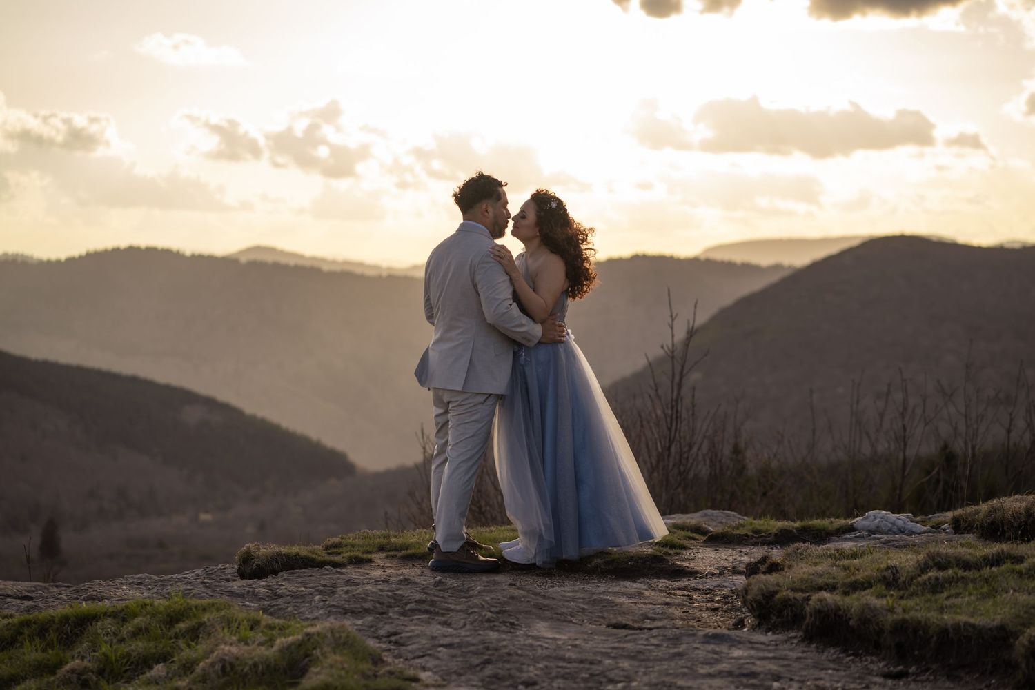 Couple in blue and white wedding attire embracing on mountain summit at golden sunset.