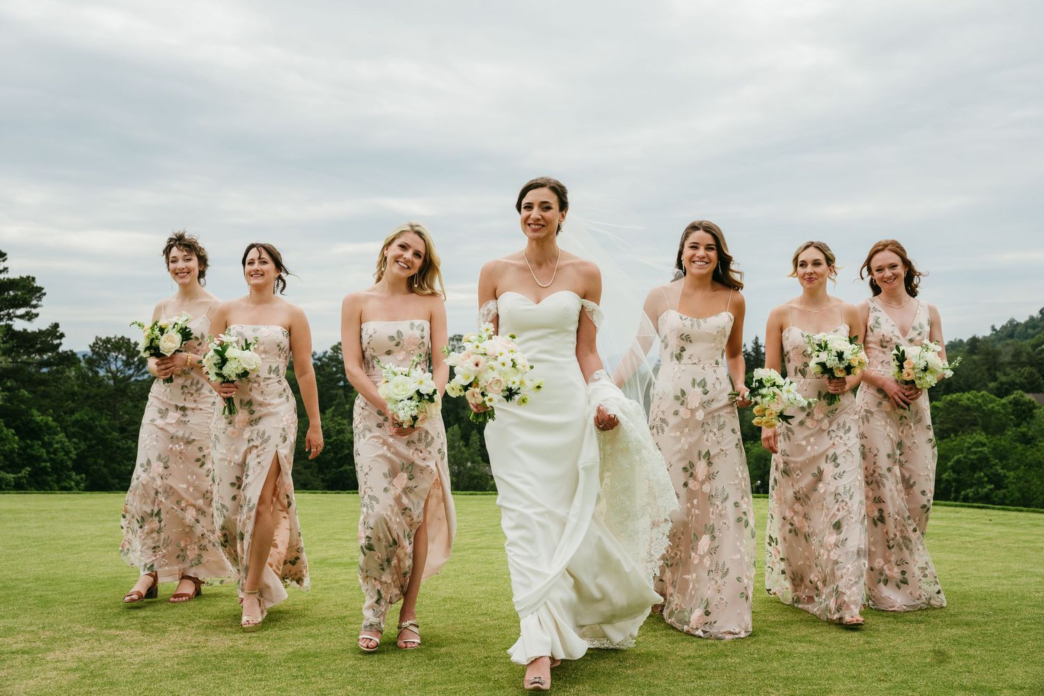 Bride and bridesmaids in floral gowns walk confidently across a green field holding white floral bouquets.