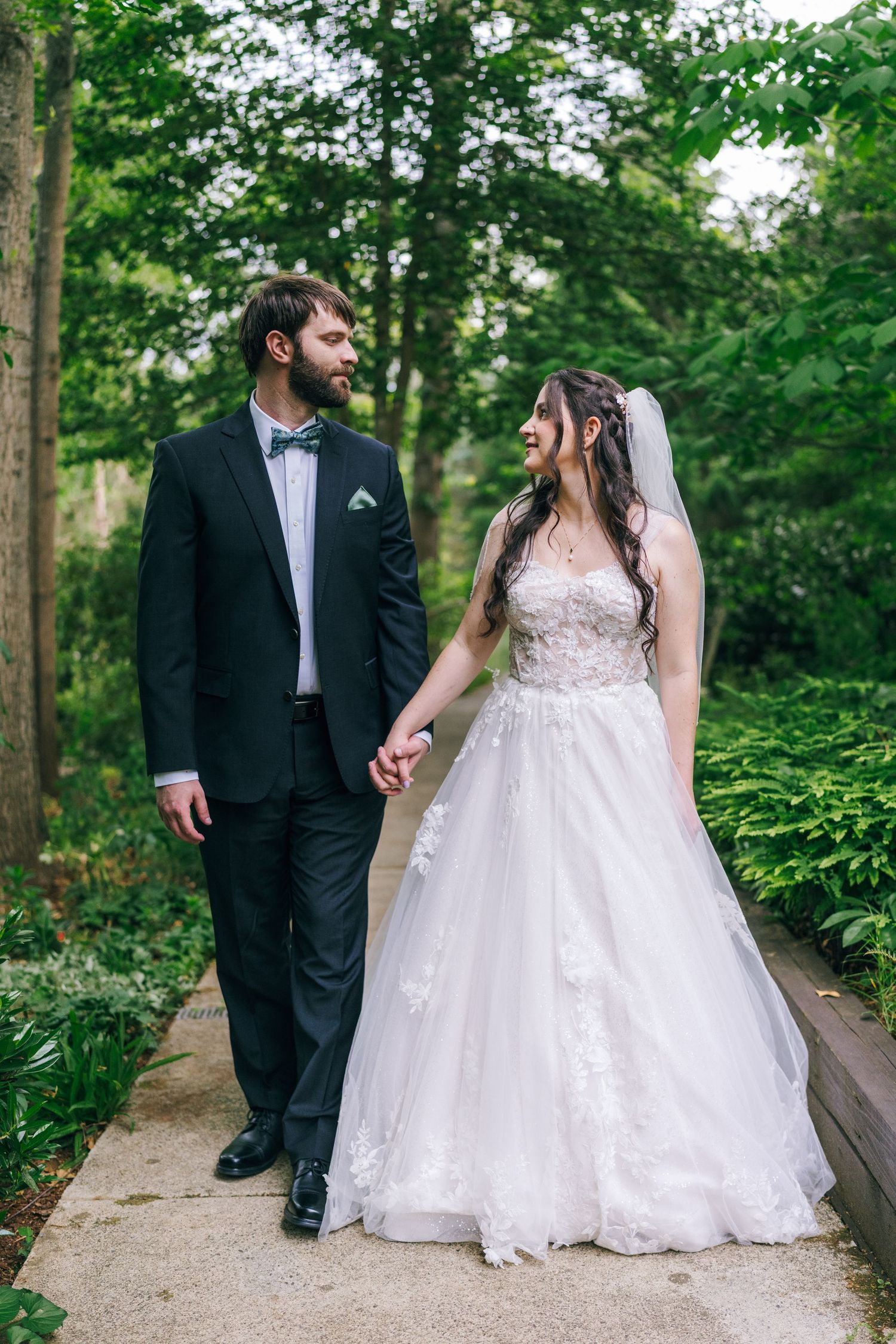 Bride and groom hold hands walking along a garden path surrounded by lush green trees.