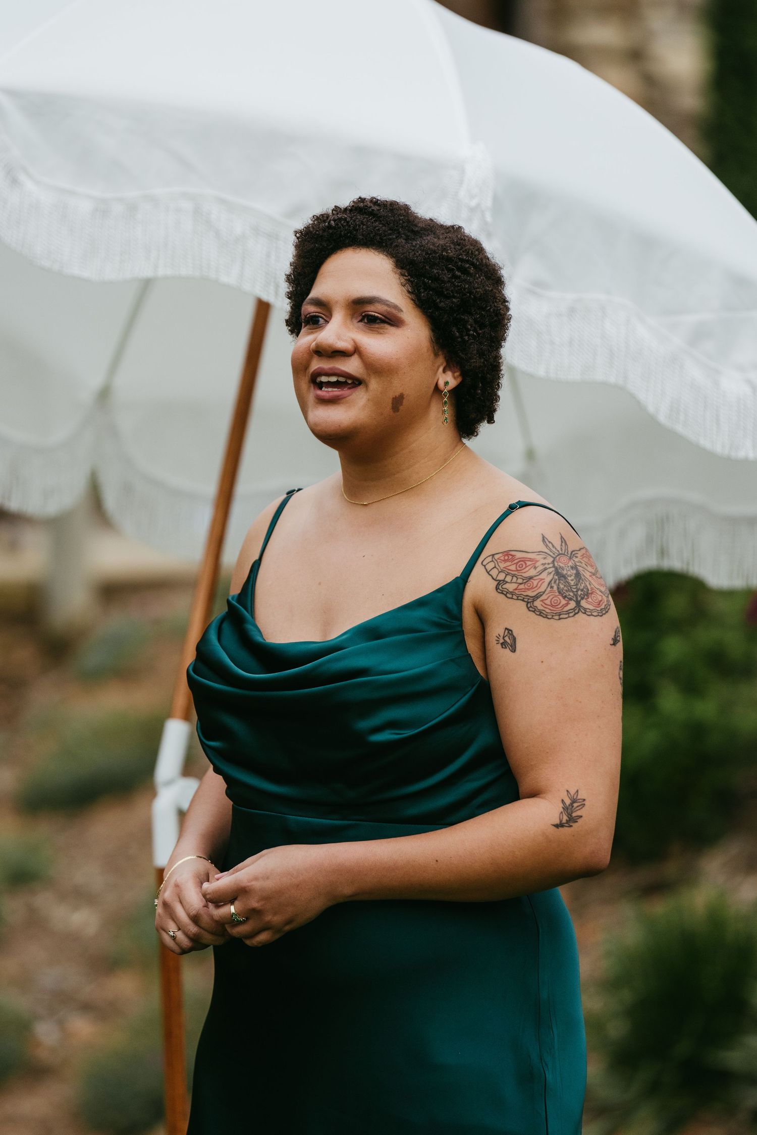A woman in a teal satin slip dress with floral tattoos stands beneath a white fringed umbrella outdoors.