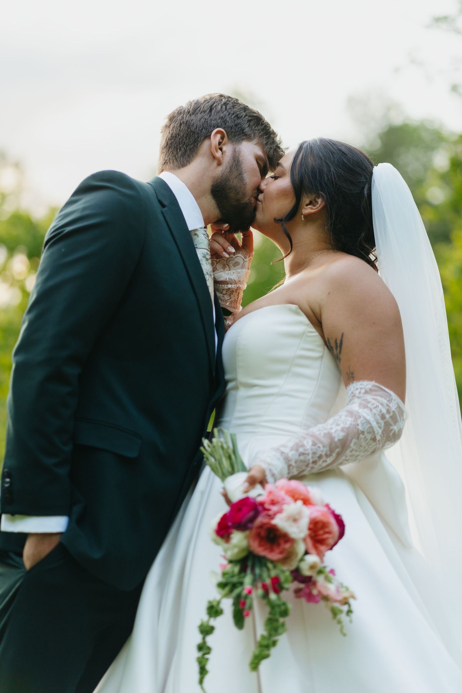 Bride and groom share a romantic kiss outdoors, bride holding a vibrant pink bouquet.
