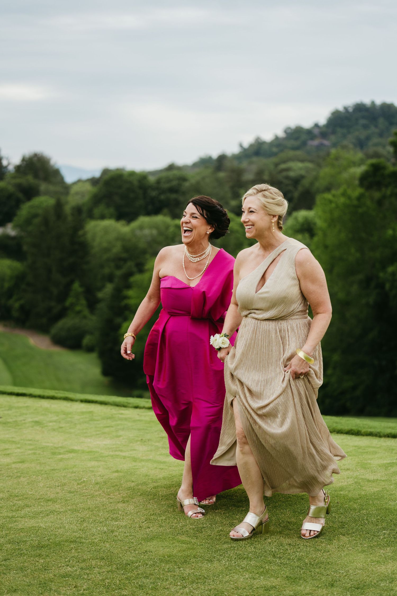 Two smiling women in formal gowns walk together across a green lawn with scenic mountain views behind them.