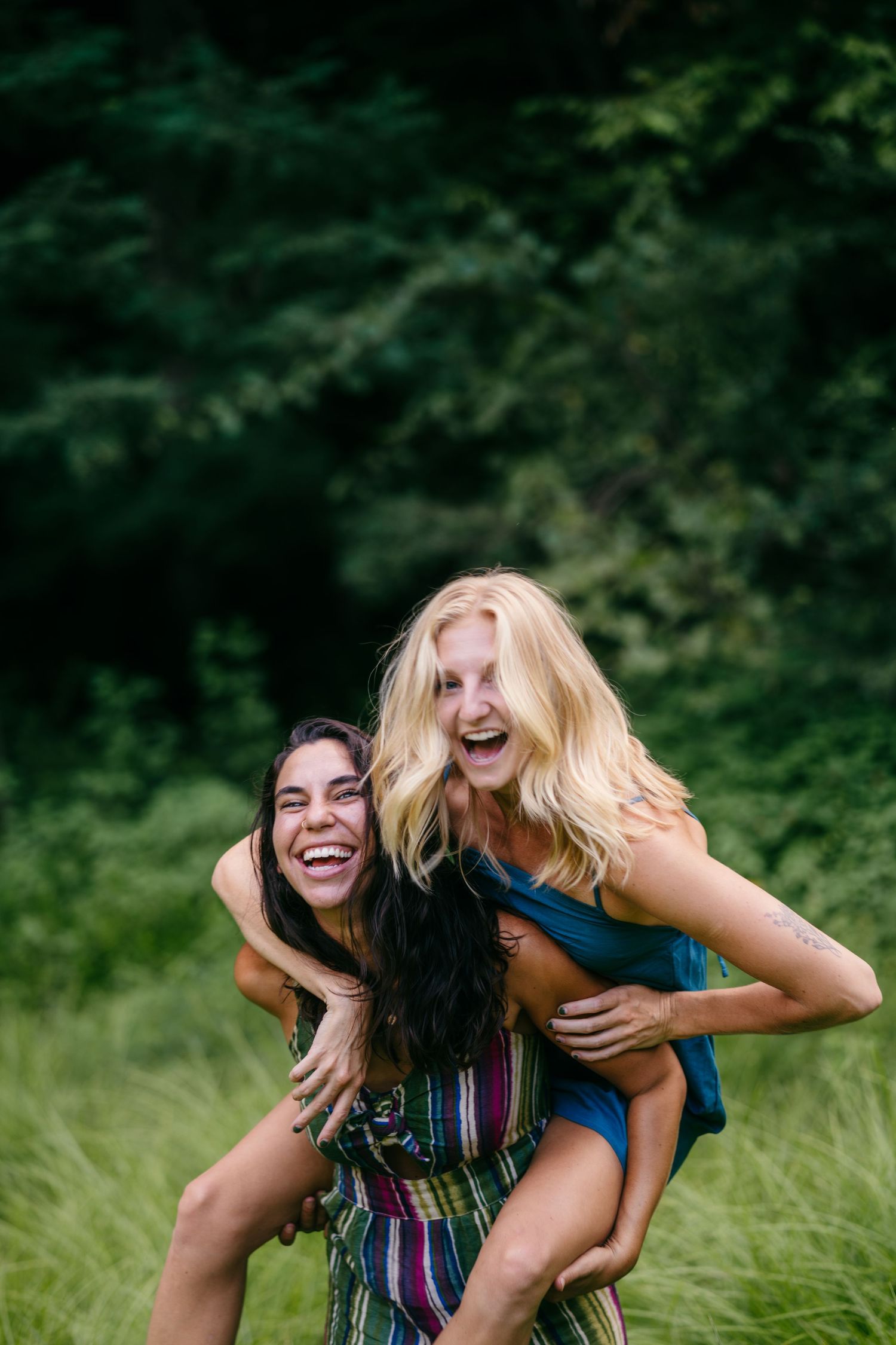Two laughing women enjoy a playful piggyback ride in a lush green outdoor setting.