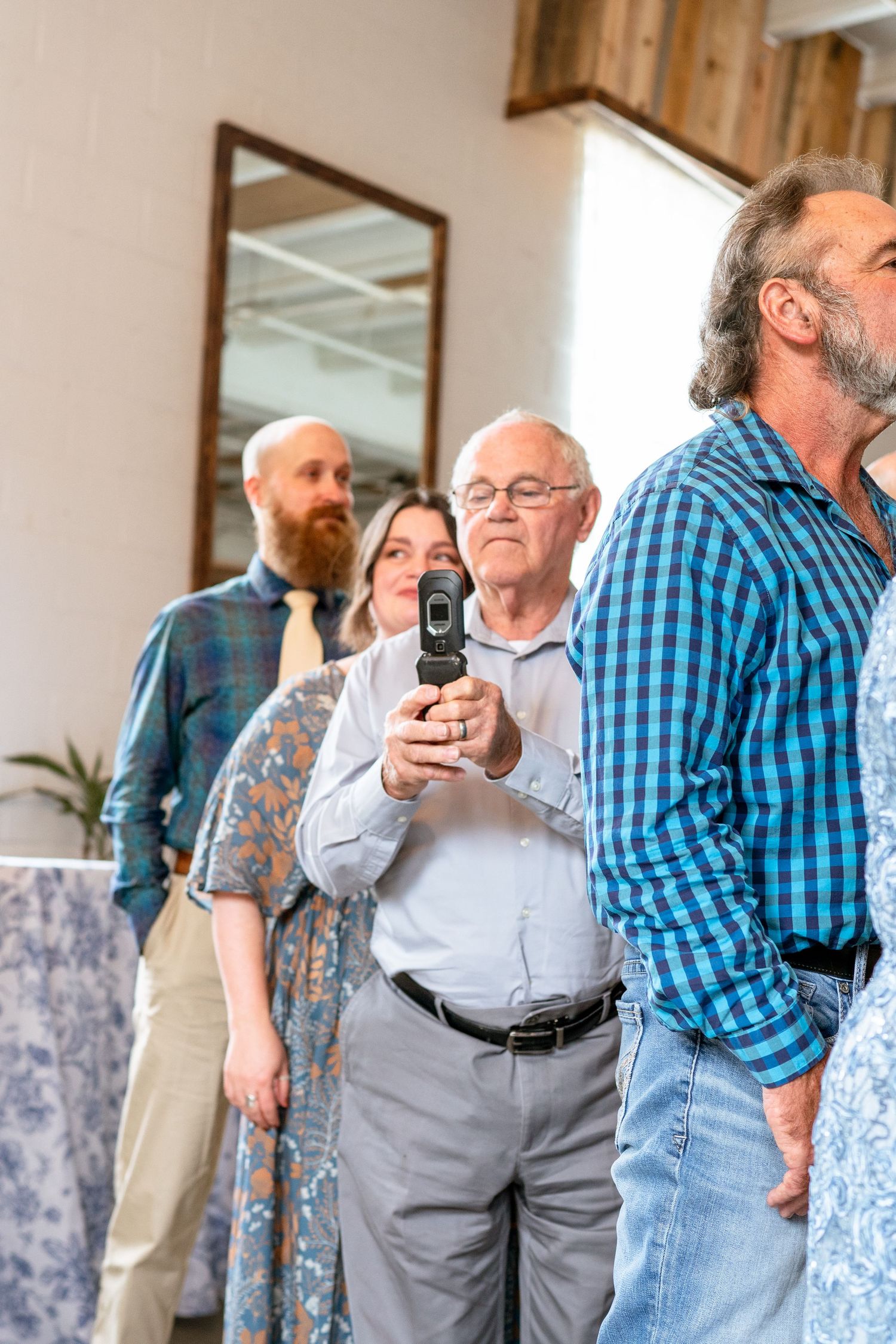Wedding guests watching ceremony, older man using flip phone to capture the moment.