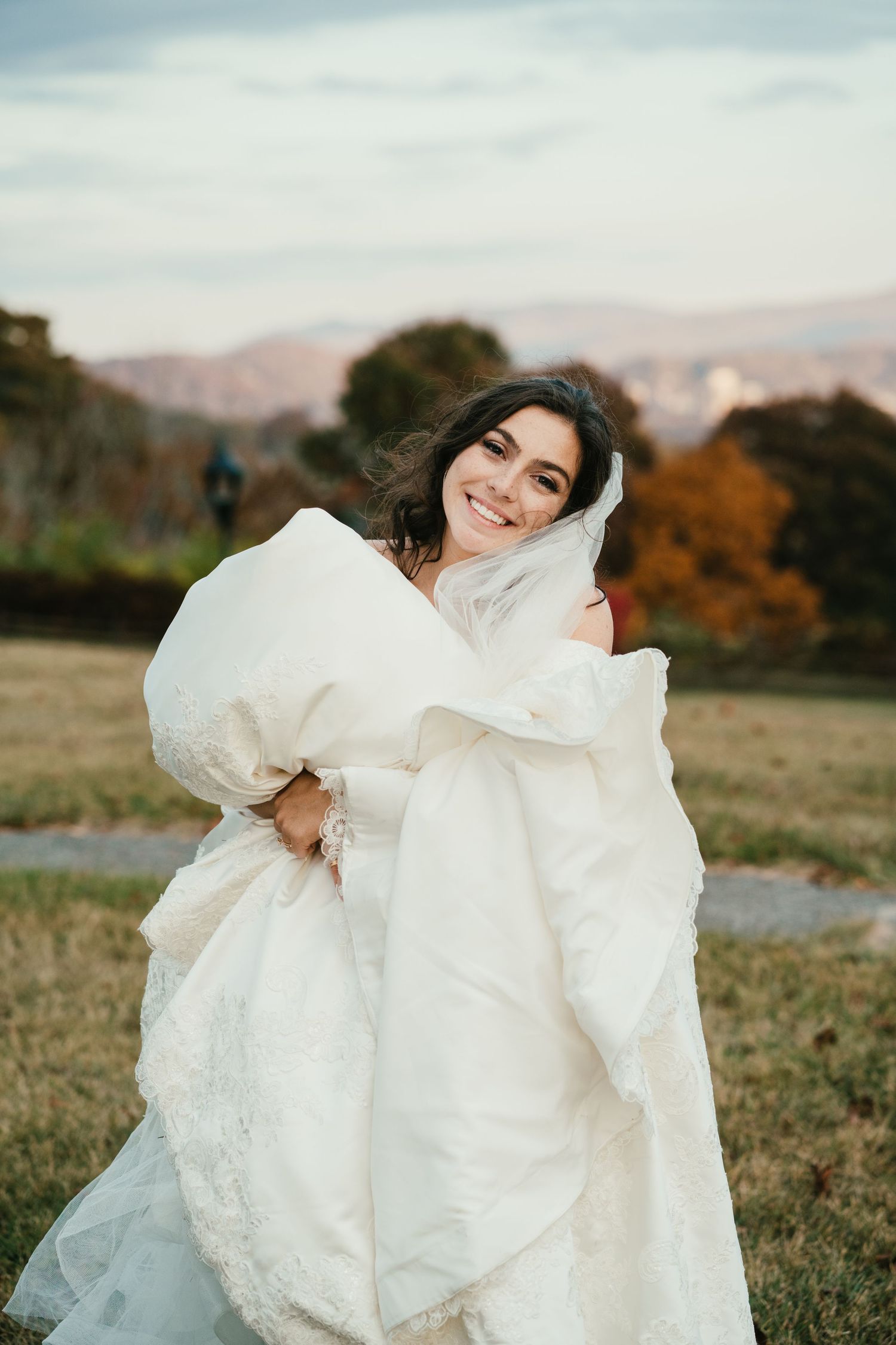 Smiling bride in a white gown holds her dress while posing outdoors with autumn mountain scenery.