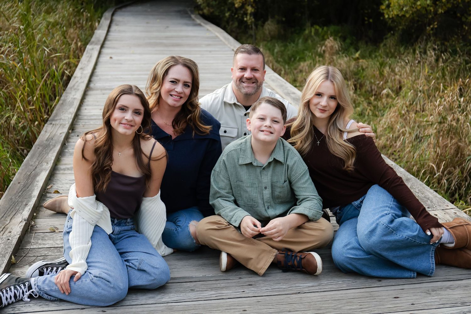 Happy family of five sitting together on wooden boardwalk surrounded by autumn foliage.