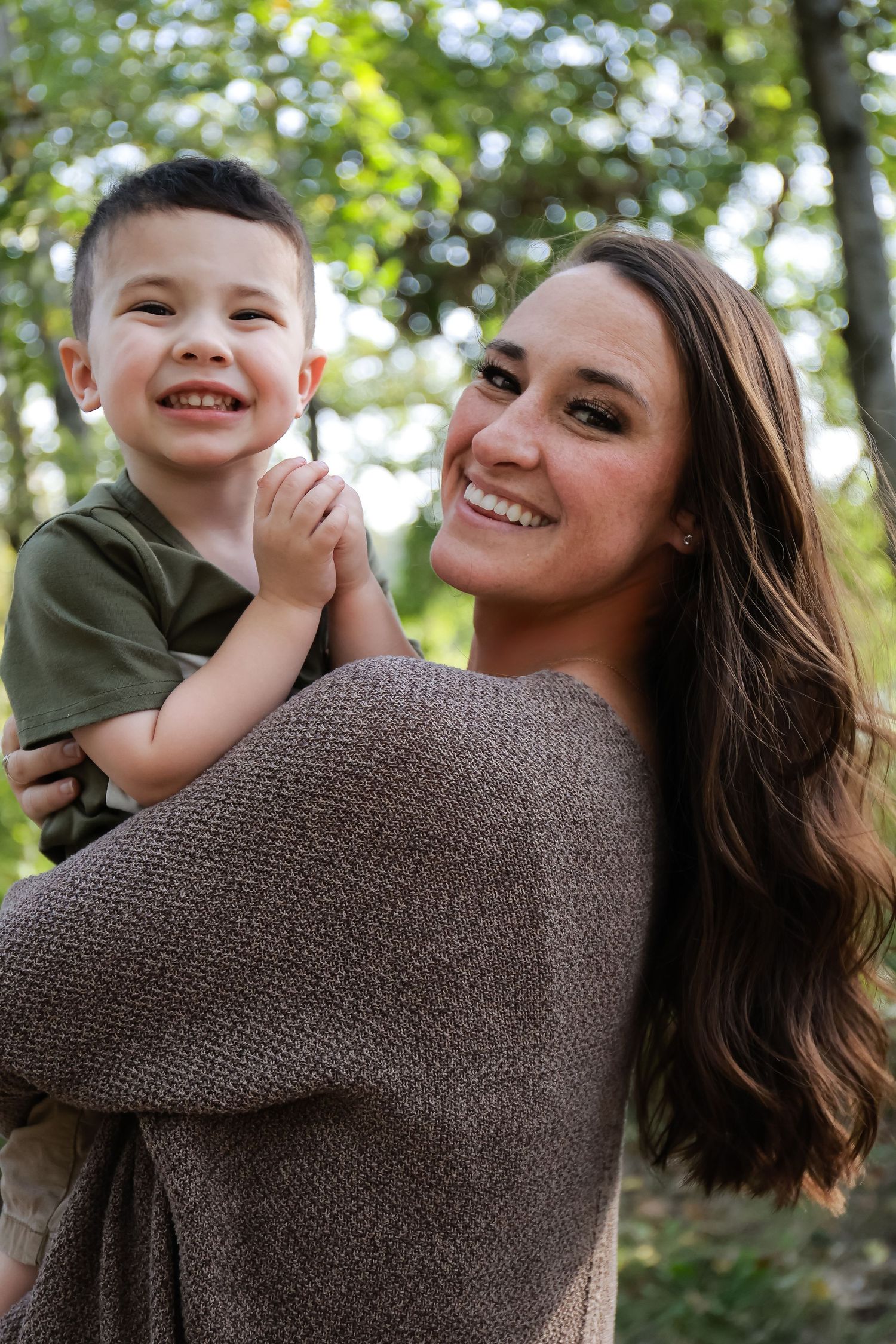Smiling mother with long dark hair holding happy young boy in green shirt in a wooded outdoor setting.