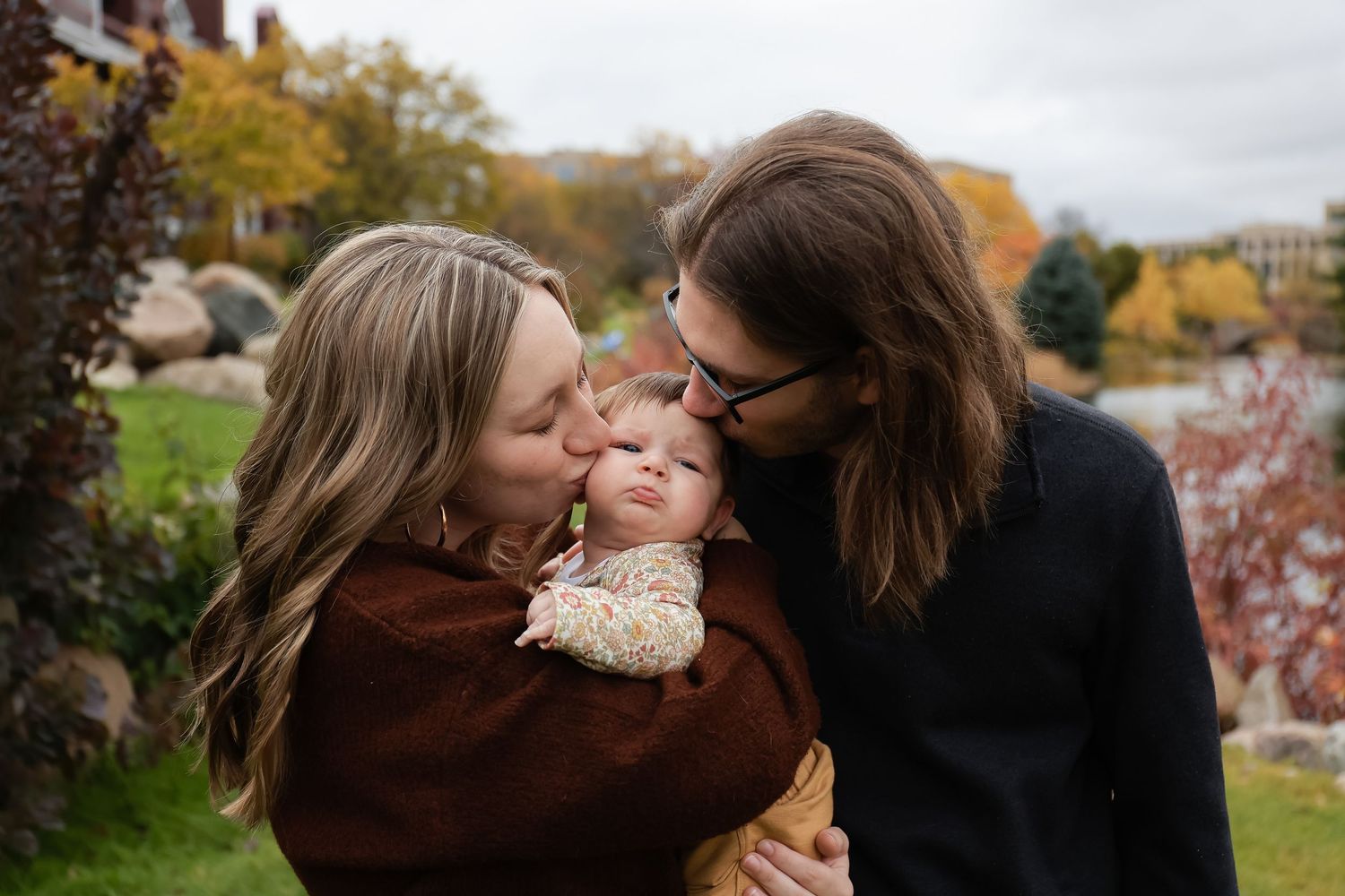 Two parents kissing their newborn baby on the cheeks in a colorful autumn outdoor setting.