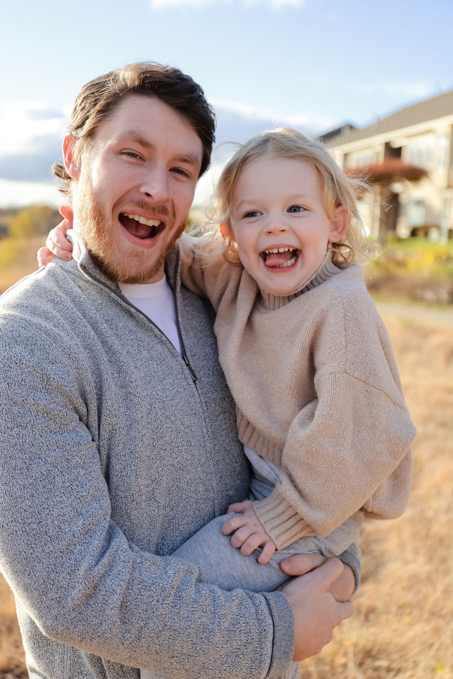 Laughing father holding smiling toddler daughter outdoors in autumn field wearing matching gray and beige sweaters.